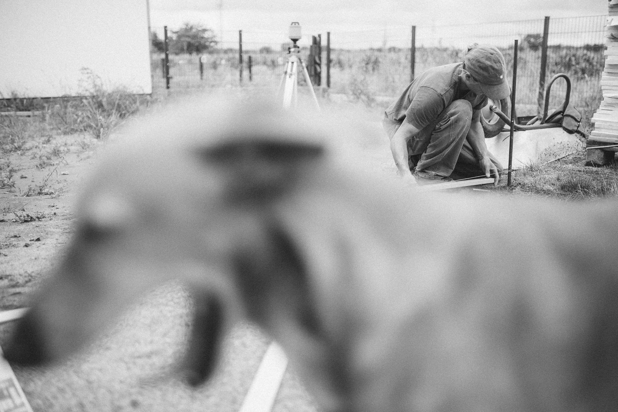 Ein Arbeiter auf einer Baustelle, der in die Erde bohrt, fotografiert aus der Nähe, unscharf im Vordergrund ein Tierkopf.