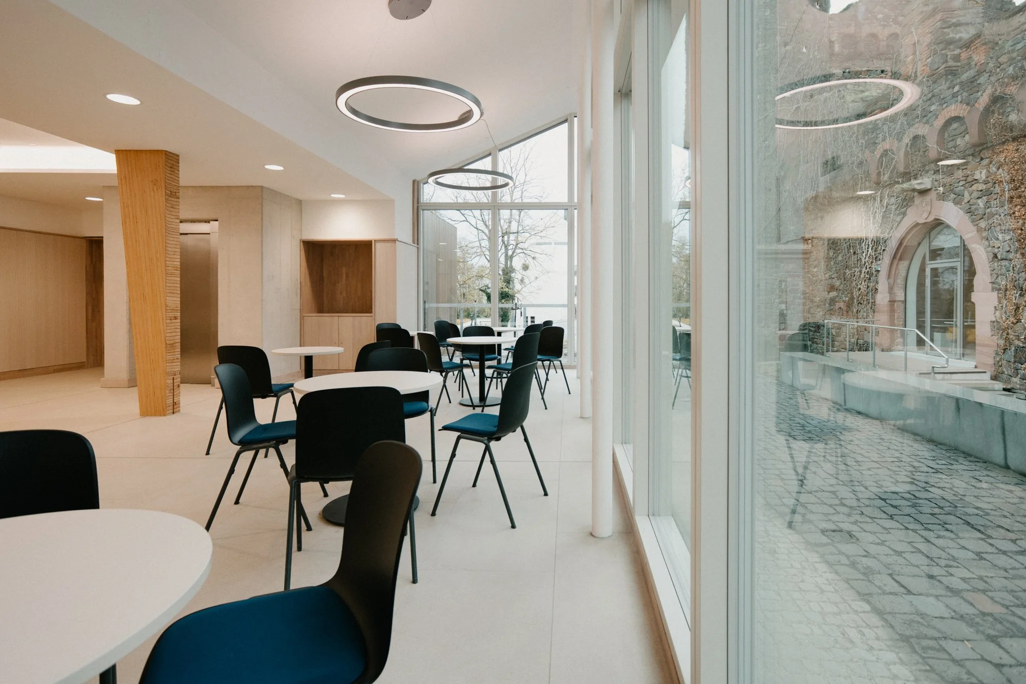 Innenraum eines modernen Cafés mit weißen Tischen, schwarzen Stühlen und großer Fensterfront, durch die man Außenbereich mit Kopfsteinpflaster und historischem Gebäude sieht.