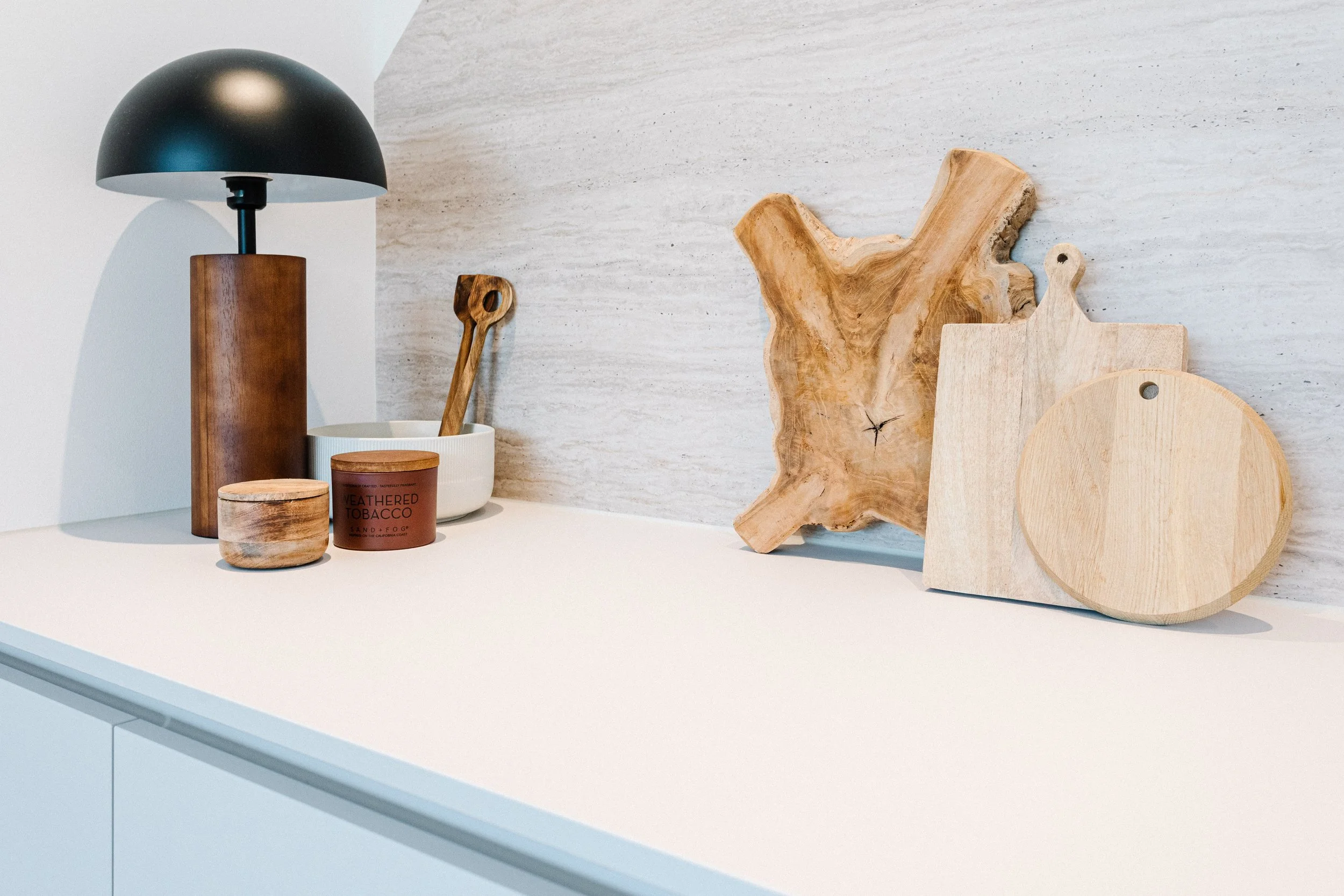 Deko auf einer weißen Kommode, bestehend aus Lampenschirm, Holzschale, Kerze, Holzlöffel, Holzschnitt, Holzscheibe und Schneidebretter gegen eine helle Wand.