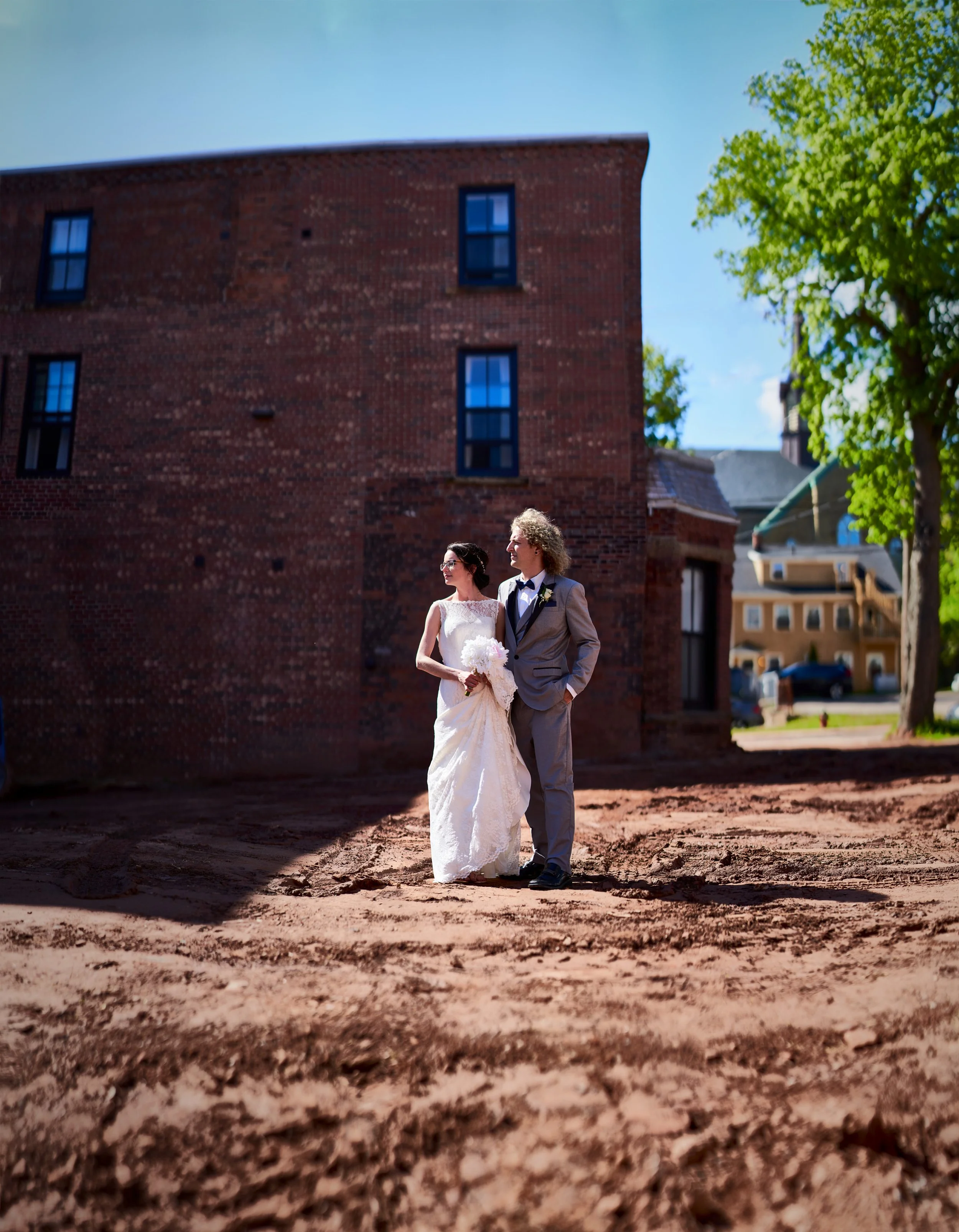 A bride and groom in wedding attire standing outside on a construction lot with a brick building behind them, trees, and a blue sky.