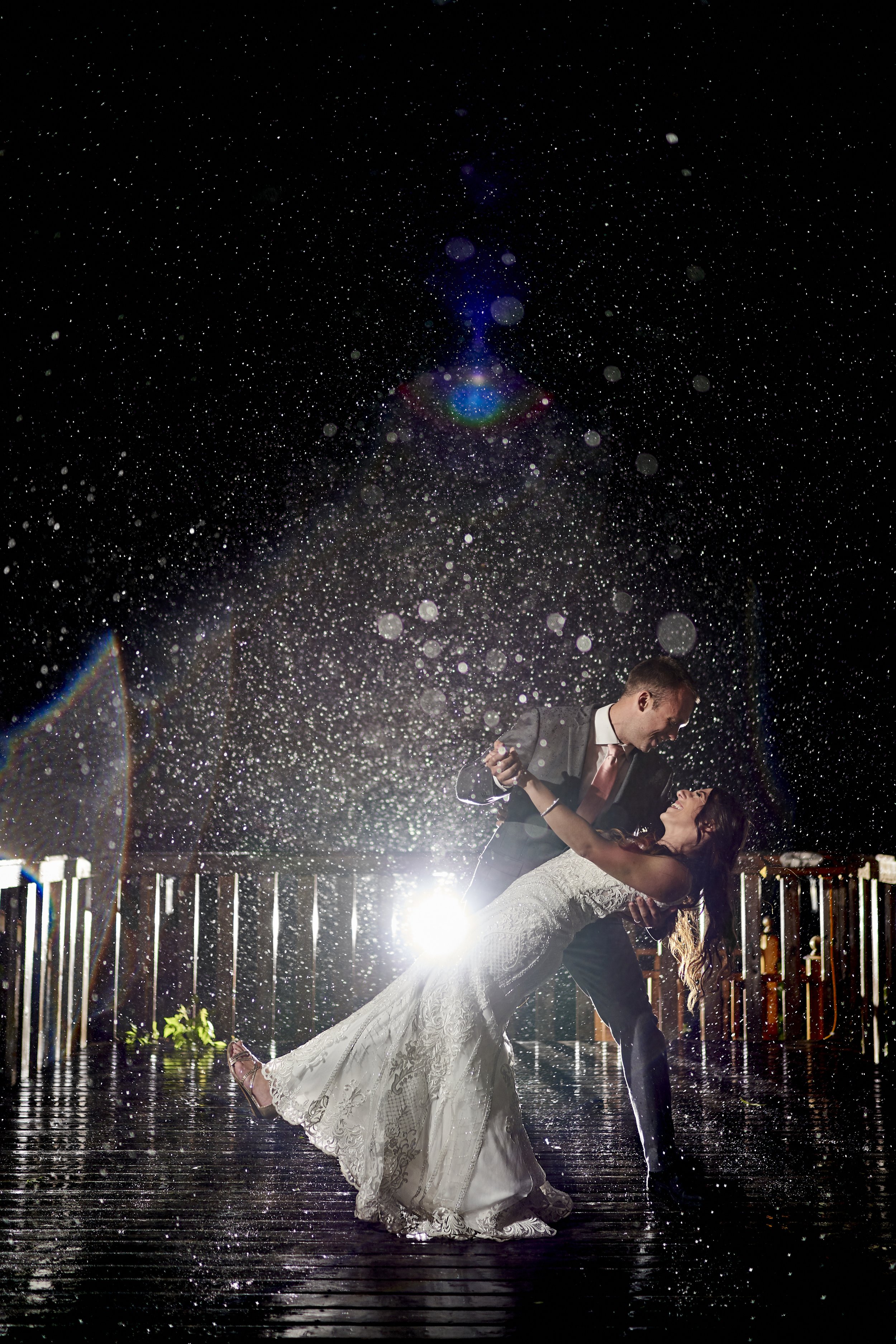 A couple in formal attire dancing on a rain-soaked outdoor deck at night with snow falling and a bright light behind them, creating a lens flare.