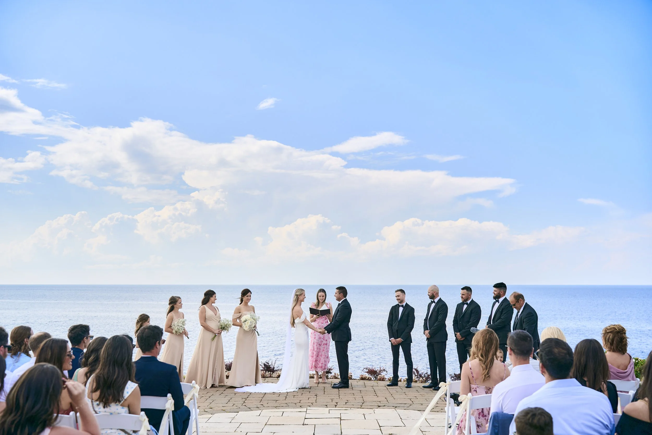 A seaside wedding ceremony with the bride and groom exchanging vows, surrounded by bridesmaids in beige dresses, groomsmen in black suits, and guests seated on white chairs, with the ocean and blue sky in the background.