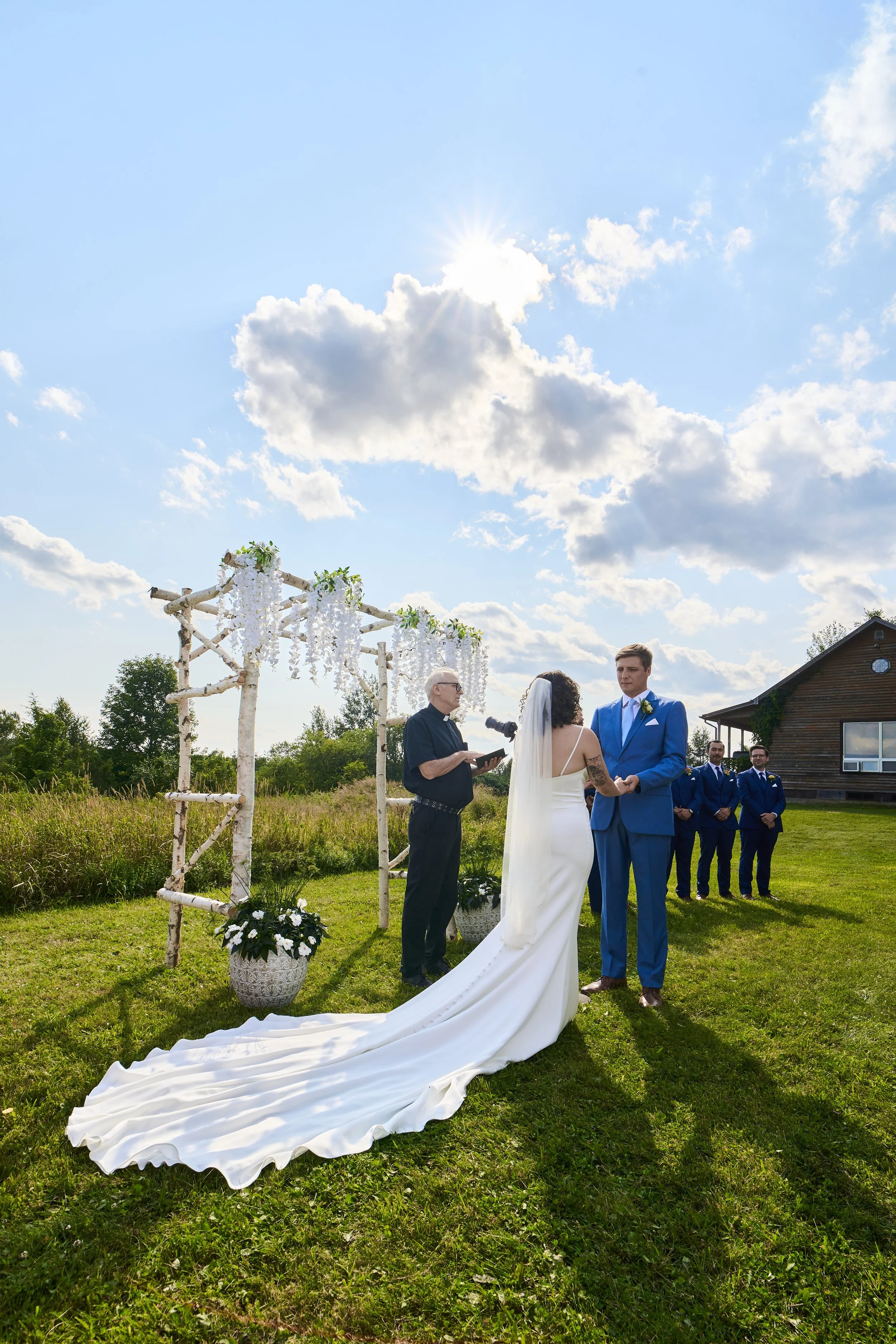 A wedding ceremony outdoors with a bride and groom standing under a decorated wooden arch, officiant, and groomsmen, on a sunny day with a blue sky and clouds.