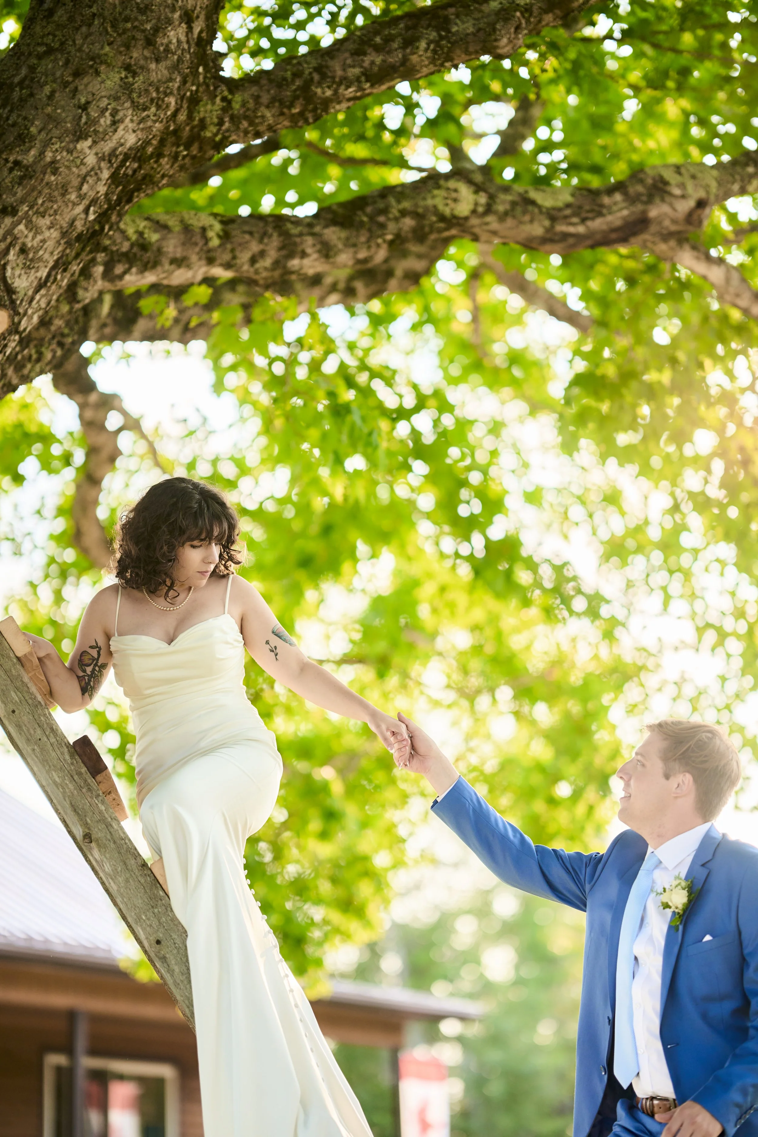 A bride in a white dress reaching out her hand to the groom, in a blue suit, outdoors on a sunny day, with green leaves and a building in the background.