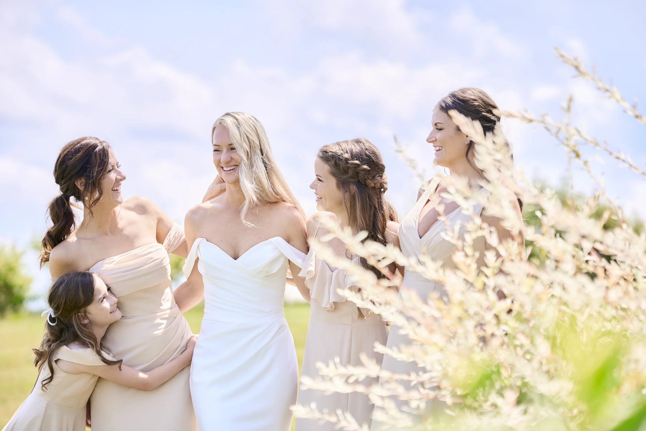 A bride and bridesmaids sharing a joyful moment outdoors on a sunny day with blue skies, surrounded by greenery and light-colored flowers.
