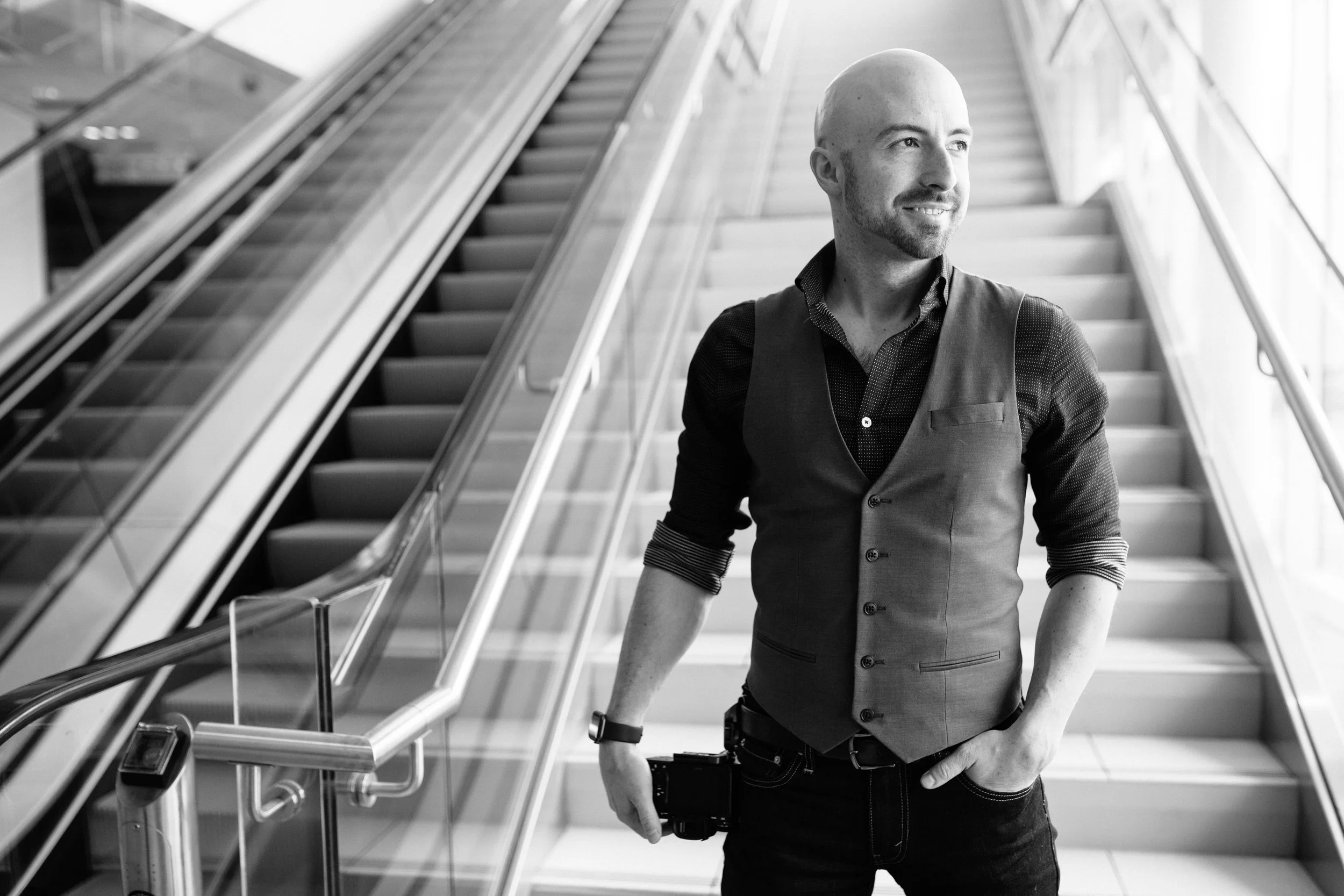 Wes Perry in a vest and shirt standing on an escalator in a modern building, holding a camera in his right hand and looking to the side.