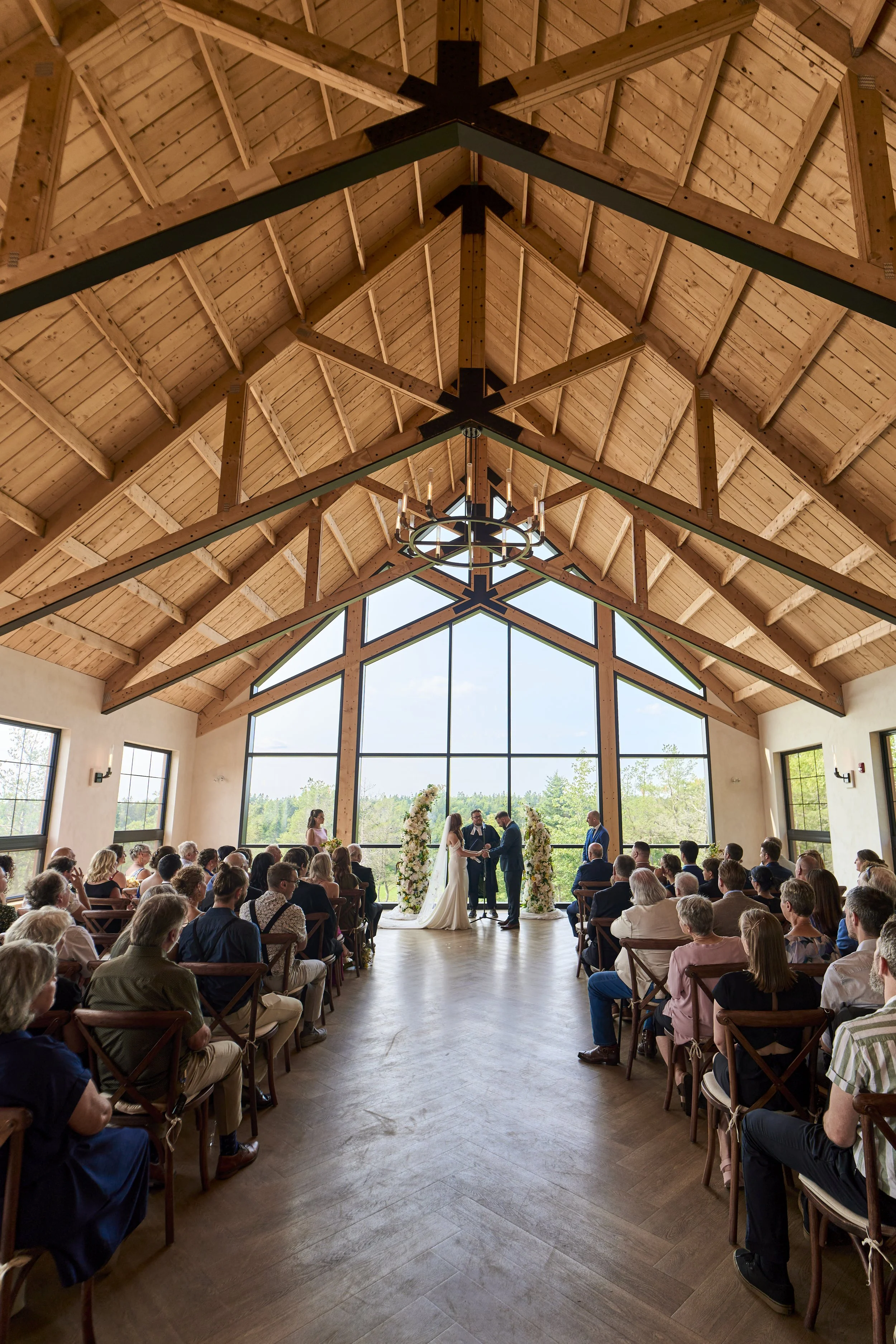 A wedding ceremony inside a spacious, rustic-style hall with a high, wooden vaulted ceiling and large glass window at the front showing an outdoor landscape. The bride and groom stand before an officiant at the altar, surrounded by floral arrangements. Guests are seated on wooden chairs on either side of the aisle, watching the ceremony.