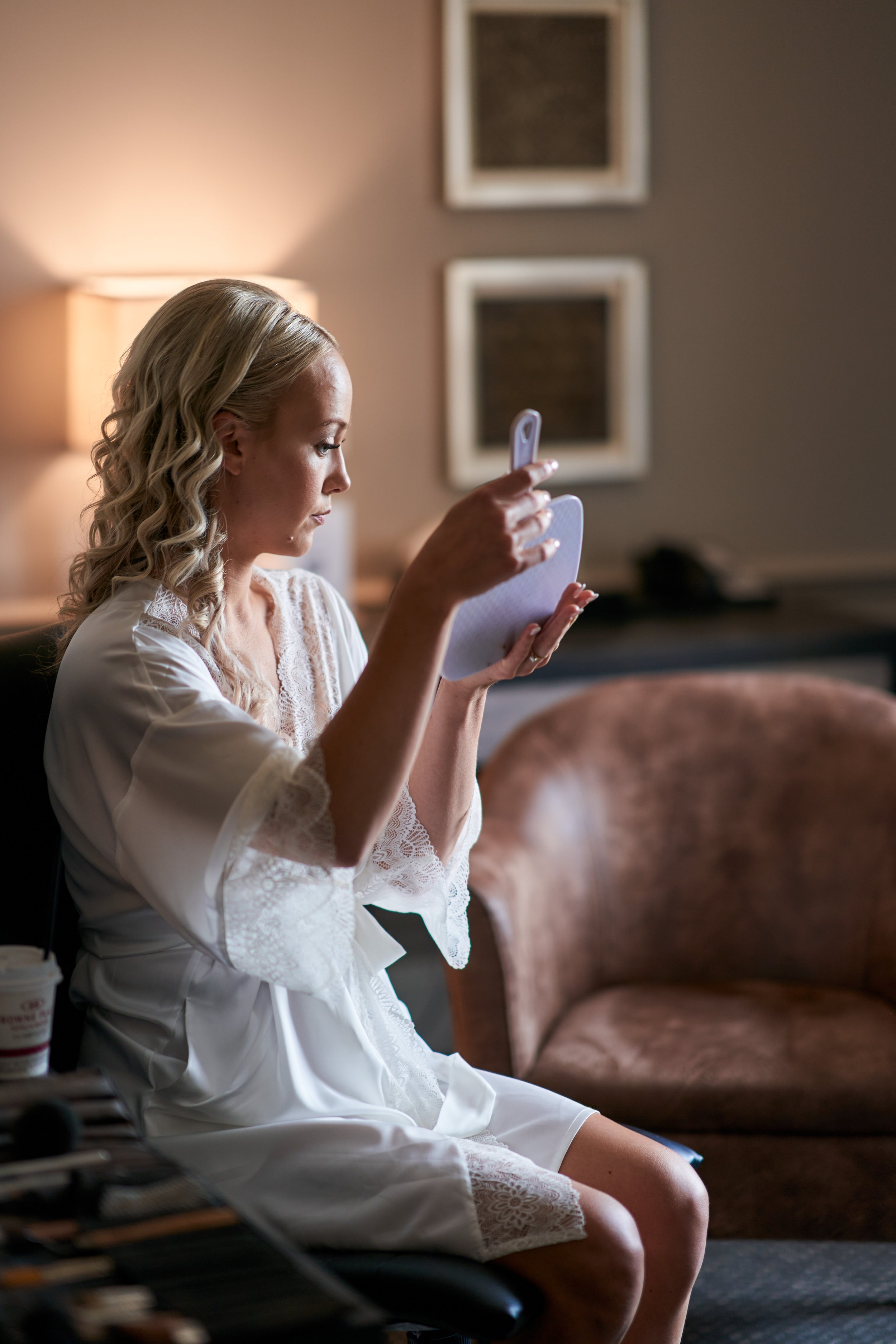 A bride with blonde curly hair in a white lace robe sitting on a chair, looking into a handheld mirror in a cozy hotel room.