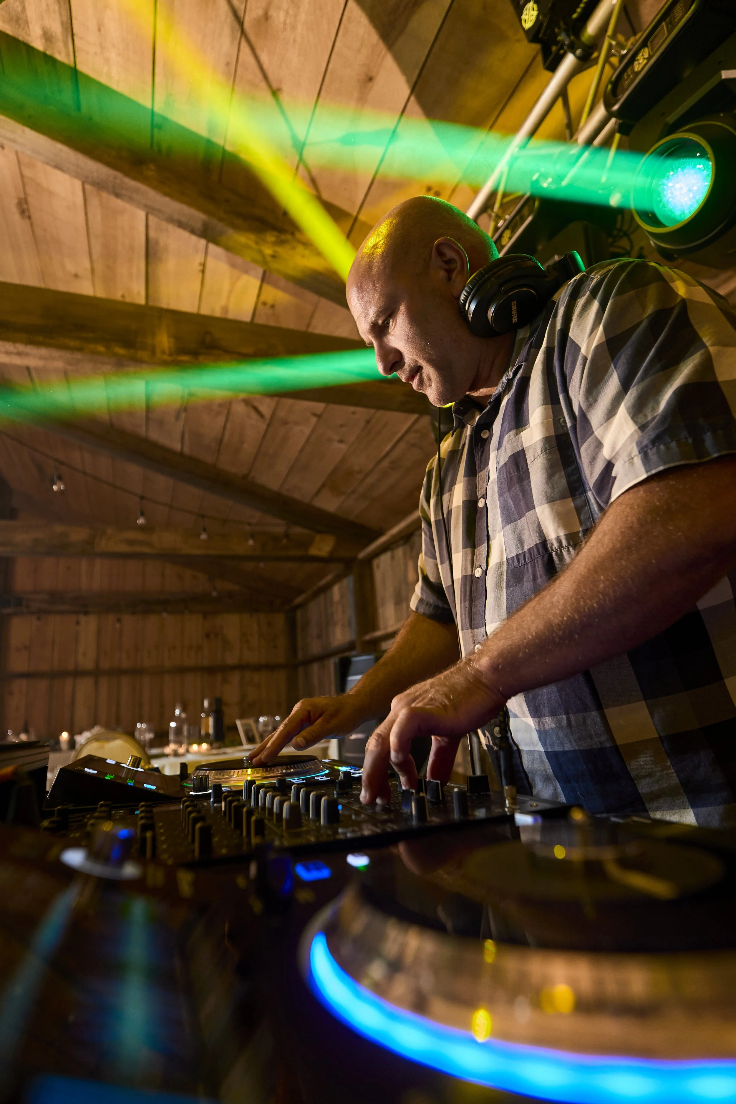 A DJ wearing headphones and a checkered shirt, working with a mixer and turntables in a wooden room with colorful lights.