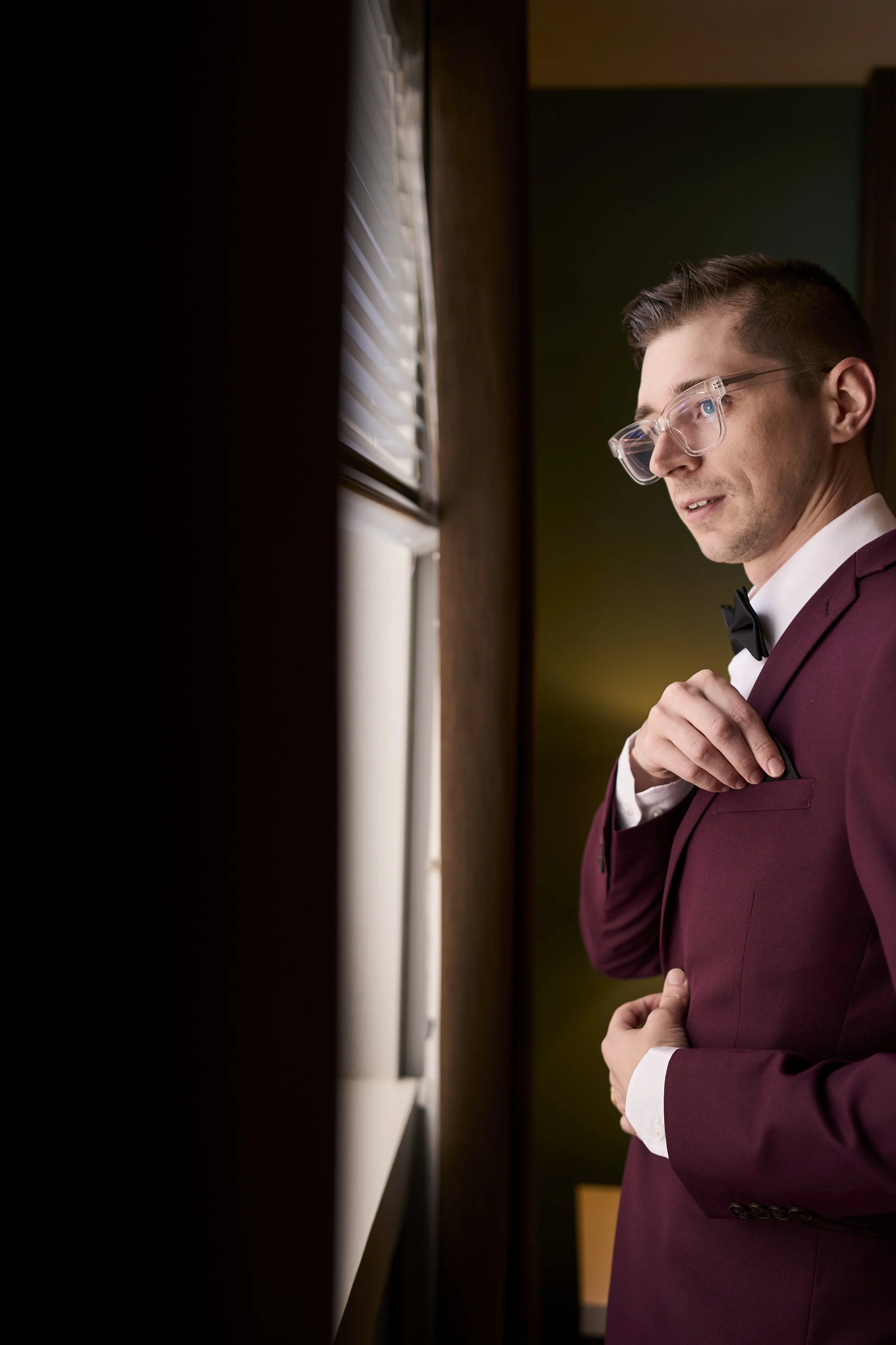 Groom in a maroon tuxedo with a black bow tie, wearing clear glasses, stands near a window adjusting his jacket.