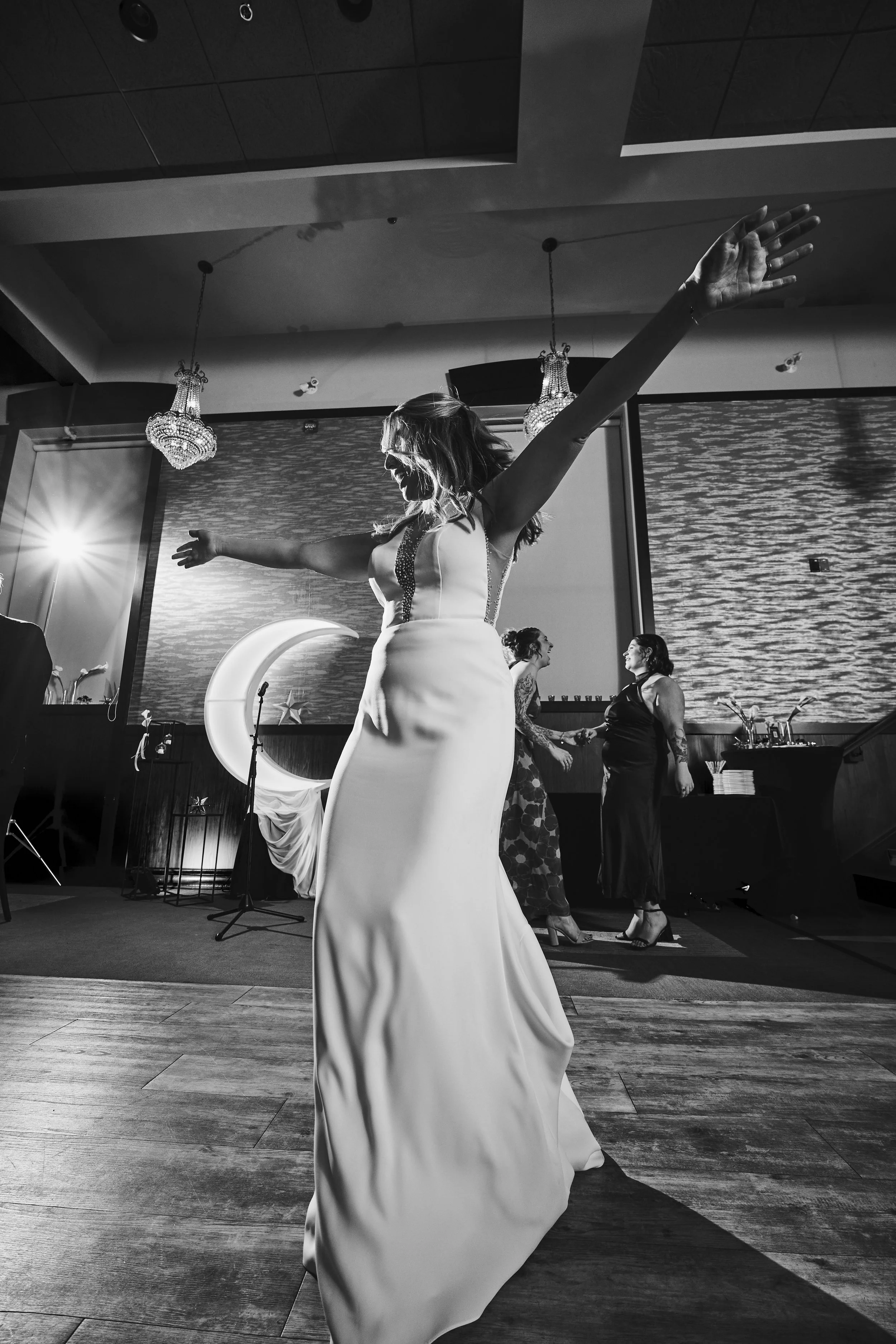 A woman in a wedding dress dancing with arms outstretched at a celebration event, with two women in the background holding hands and smiling, decorated with chandeliers and a moon-shaped decoration.