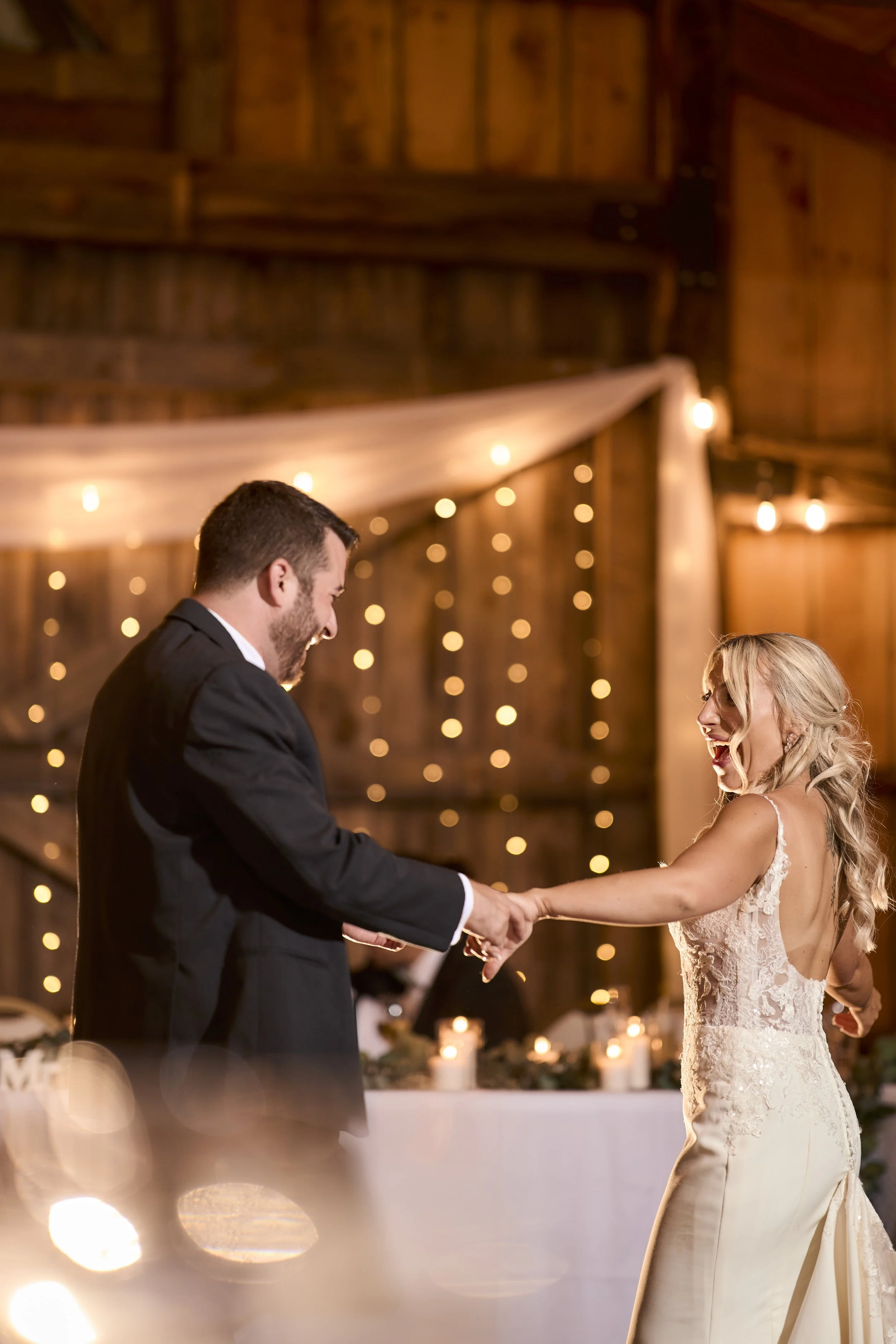 A bride and groom are dancing and smiling in a warmly lit, rustic barn wedding reception decorated with string lights and candles.