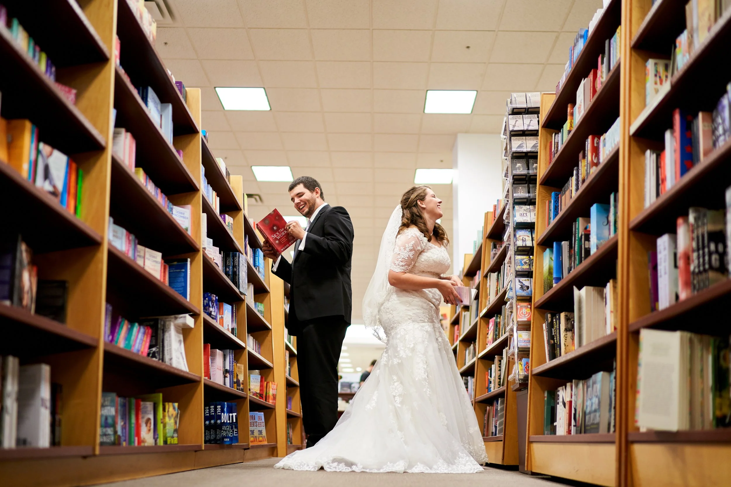 A bride and groom in wedding attire reading books in a library and enjoying each other's company.