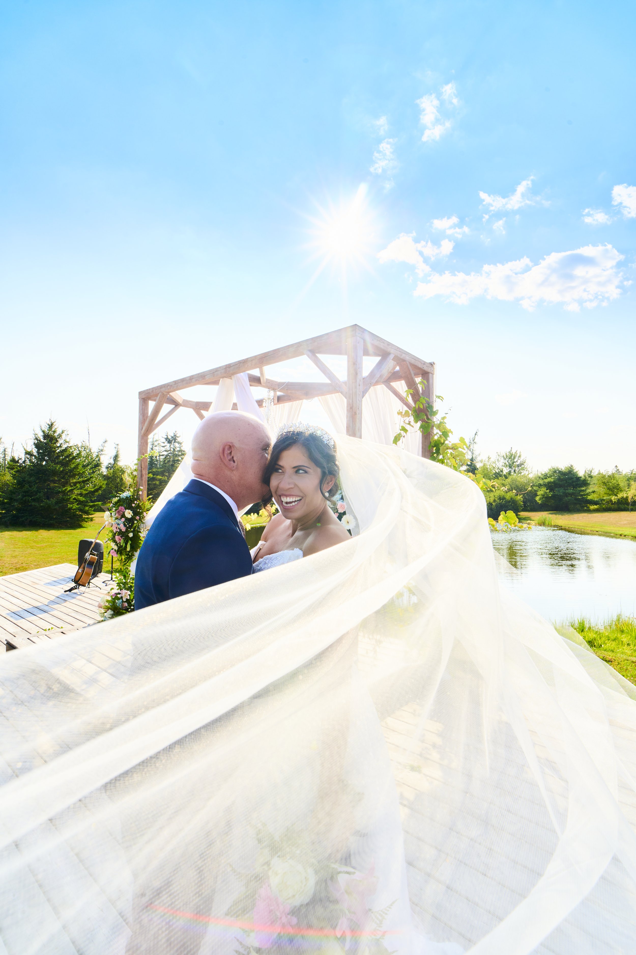 A bride and an older groom share a joyful moment outdoors during a wedding, with the bride's veil flowing in the breeze, under a sunny sky near a small pond and wooden gazebo.