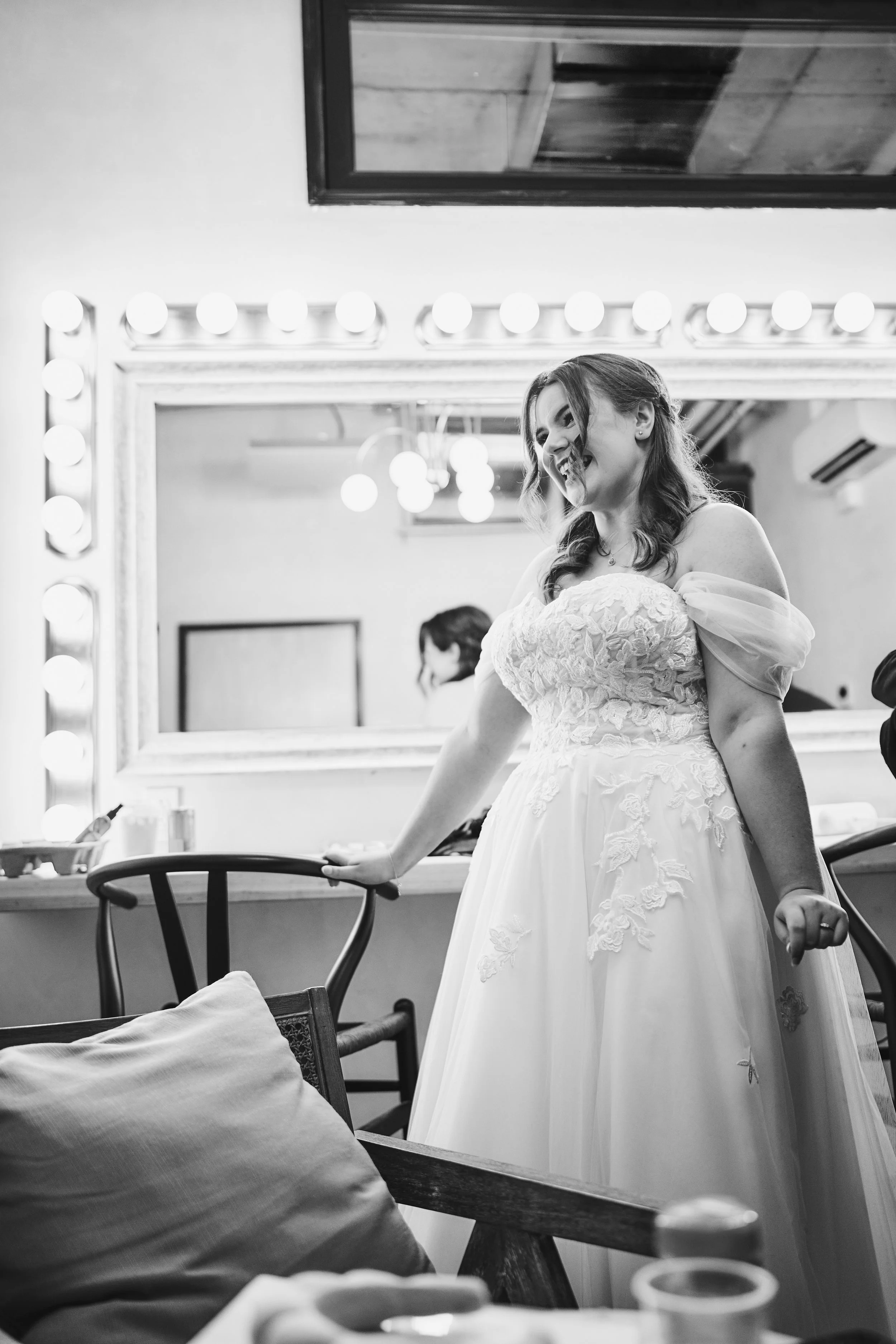 A bride in a wedding dress smiling while standing in a makeup or dressing room, with a large illuminated mirror and a person in the background.