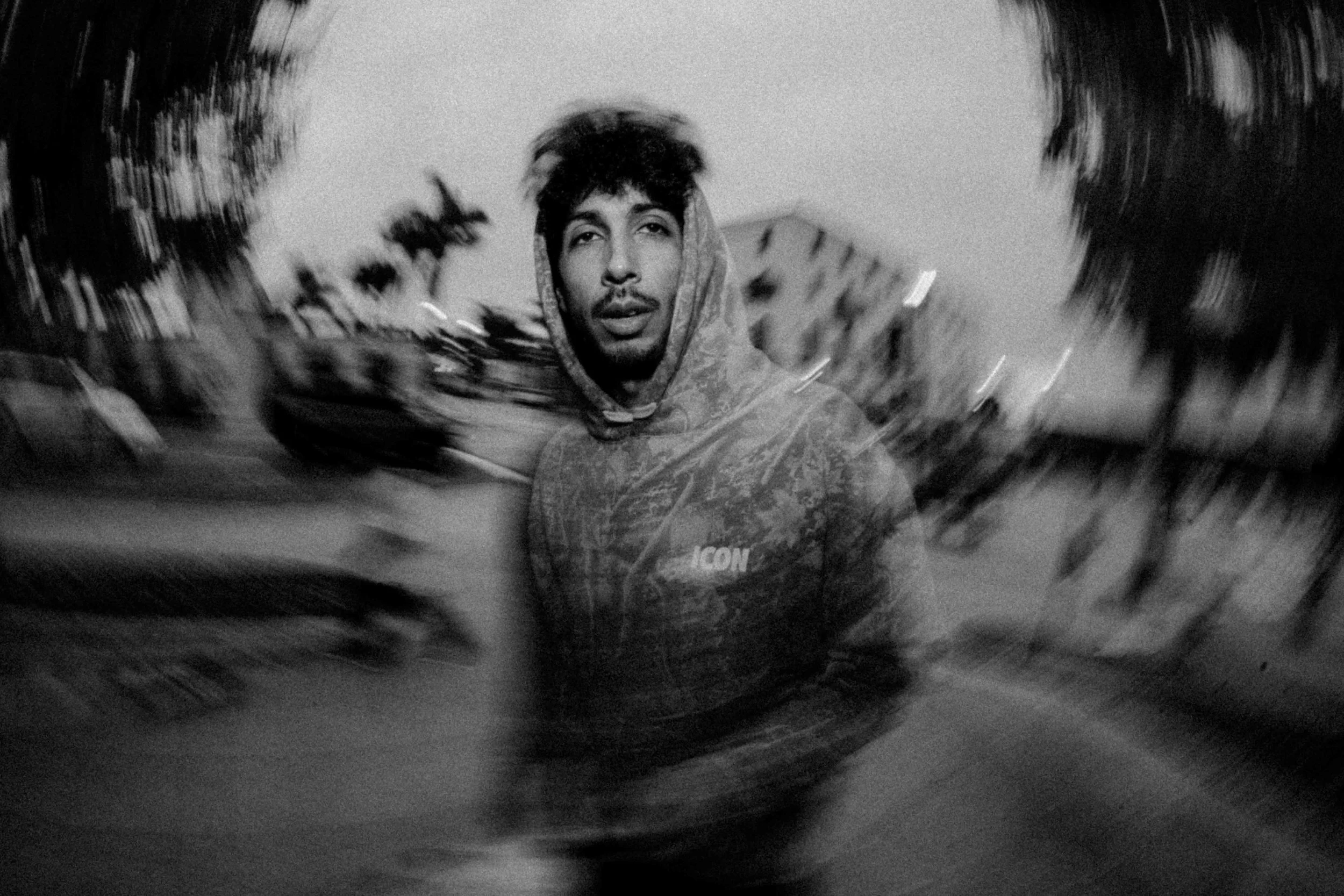 Black and white photo of a young man with dark hair and a mustache, wearing a hoodie with the word 'ICON' on it, standing outdoors with blurred trees and buildings in the background.