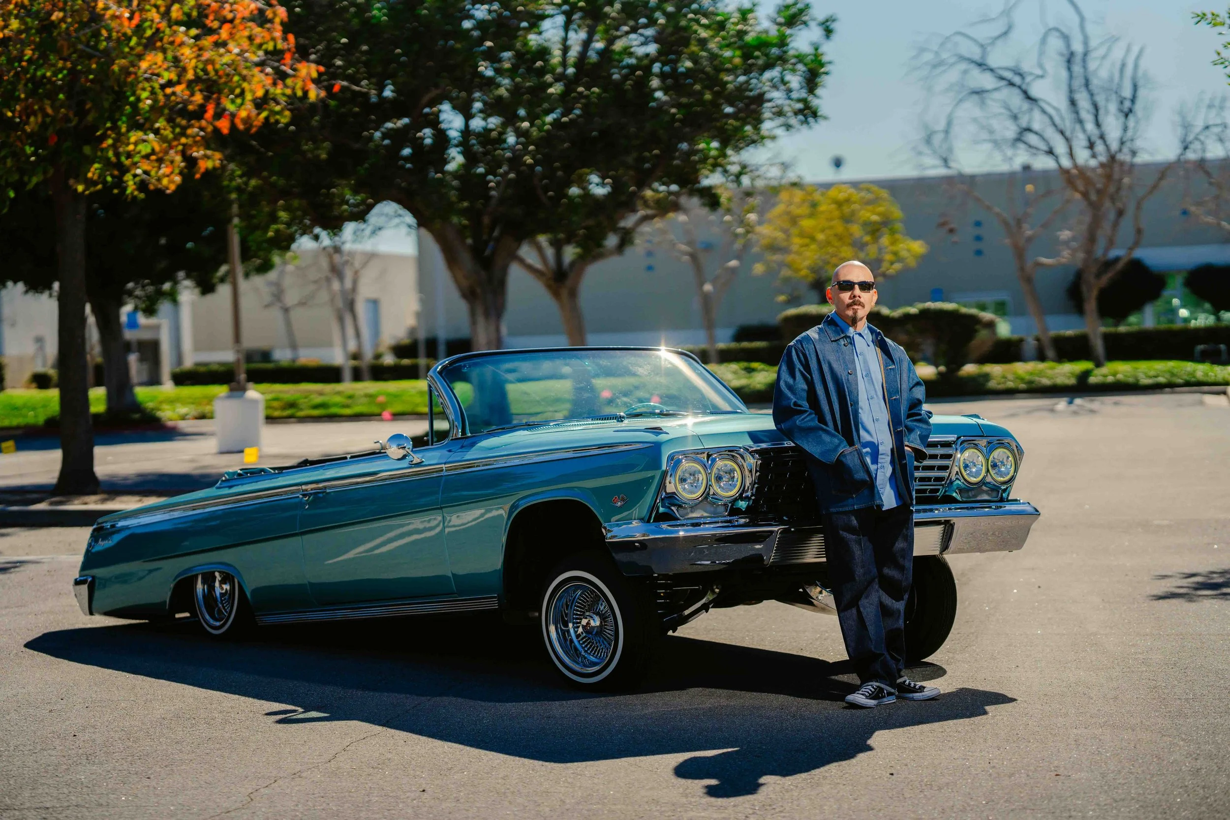 A man wearing sunglasses, a denim jacket, and dark jeans standing beside a classic blue lowrider car in an empty parking lot with trees and buildings in the background.