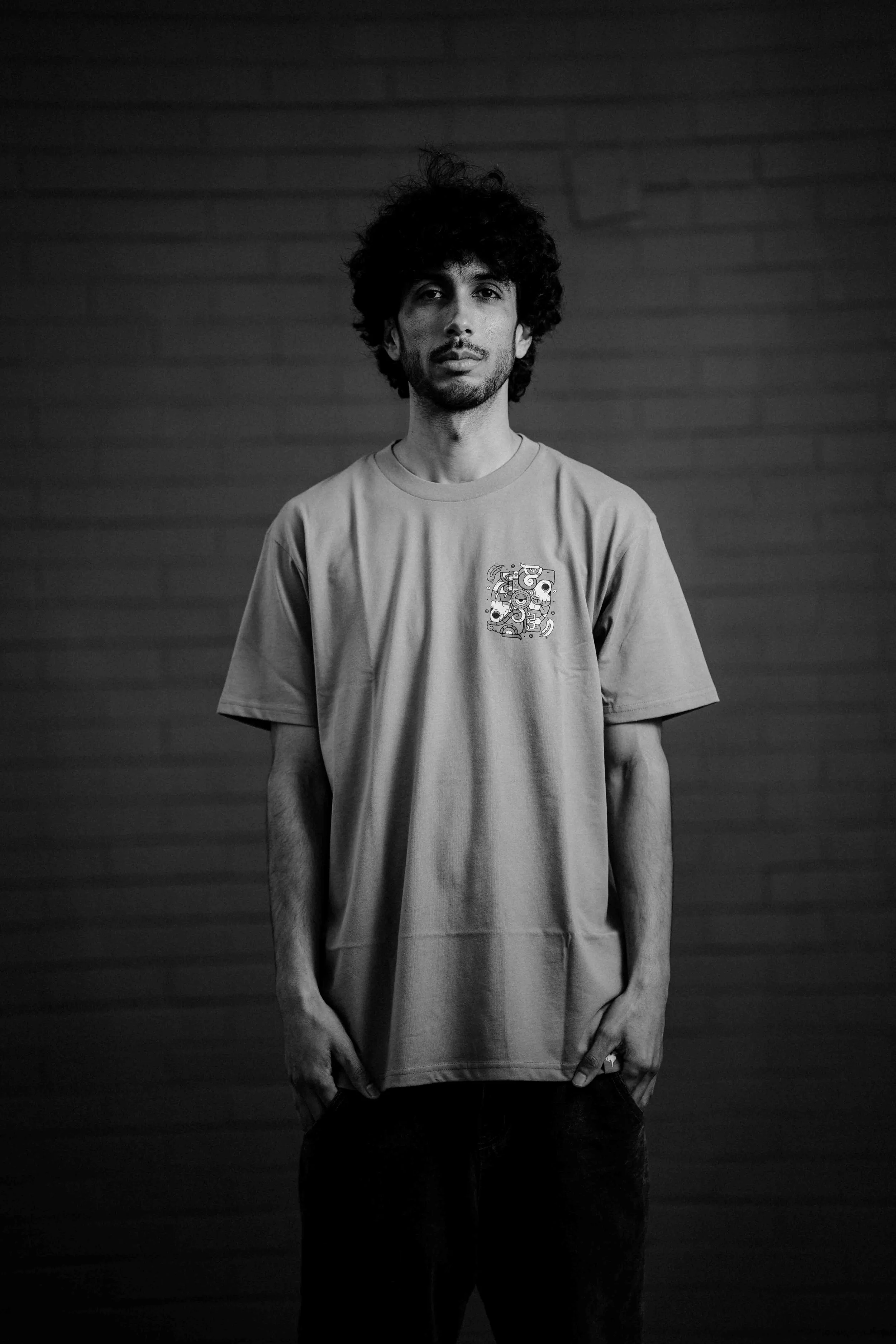 Black and white portrait of a young man with curly hair wearing a T-shirt with a small graphic on the chest, standing against a brick wall background.
