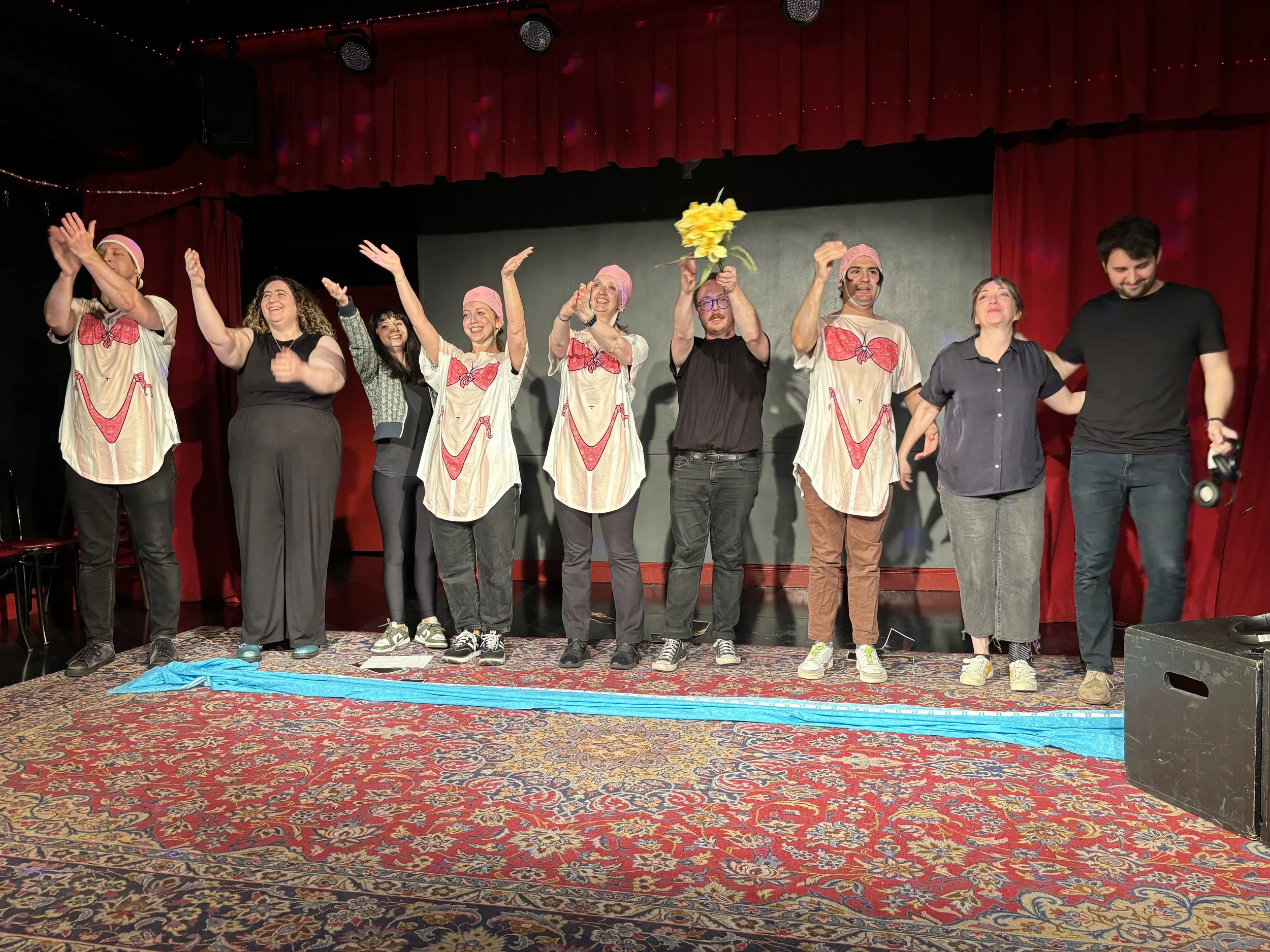 A group of nine people on stage, some dressed in pajamas with a pink sleep cap, smiling and celebrating, with a colorful patterned carpet on the floor and a red curtain backdrop.