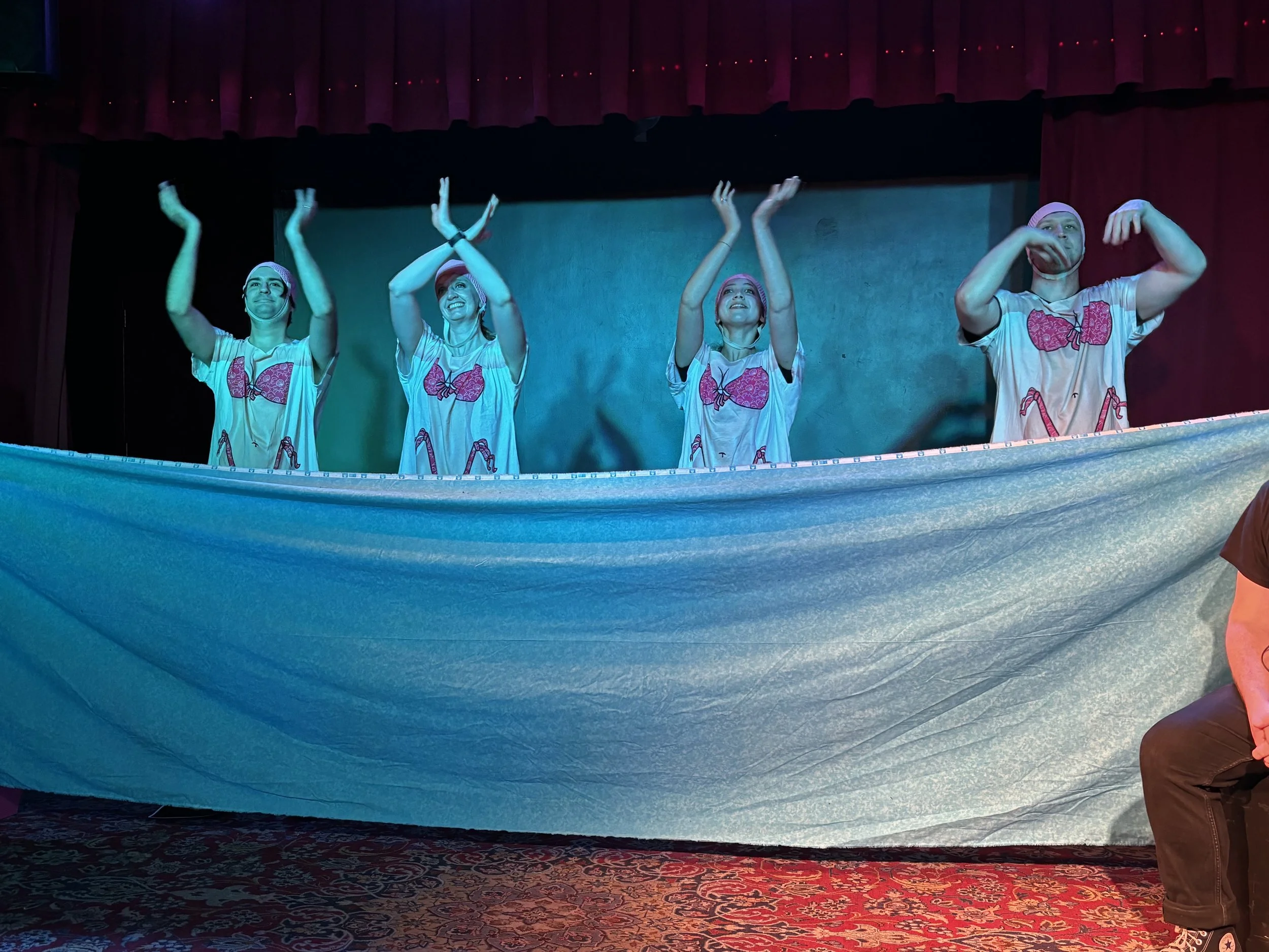 Four performers on stage wearing matching pajamas with pink hearts, pink headbands, and athletic wristbands, performing a dance with their arms raised, in front of a blue backdrop and red curtains.
