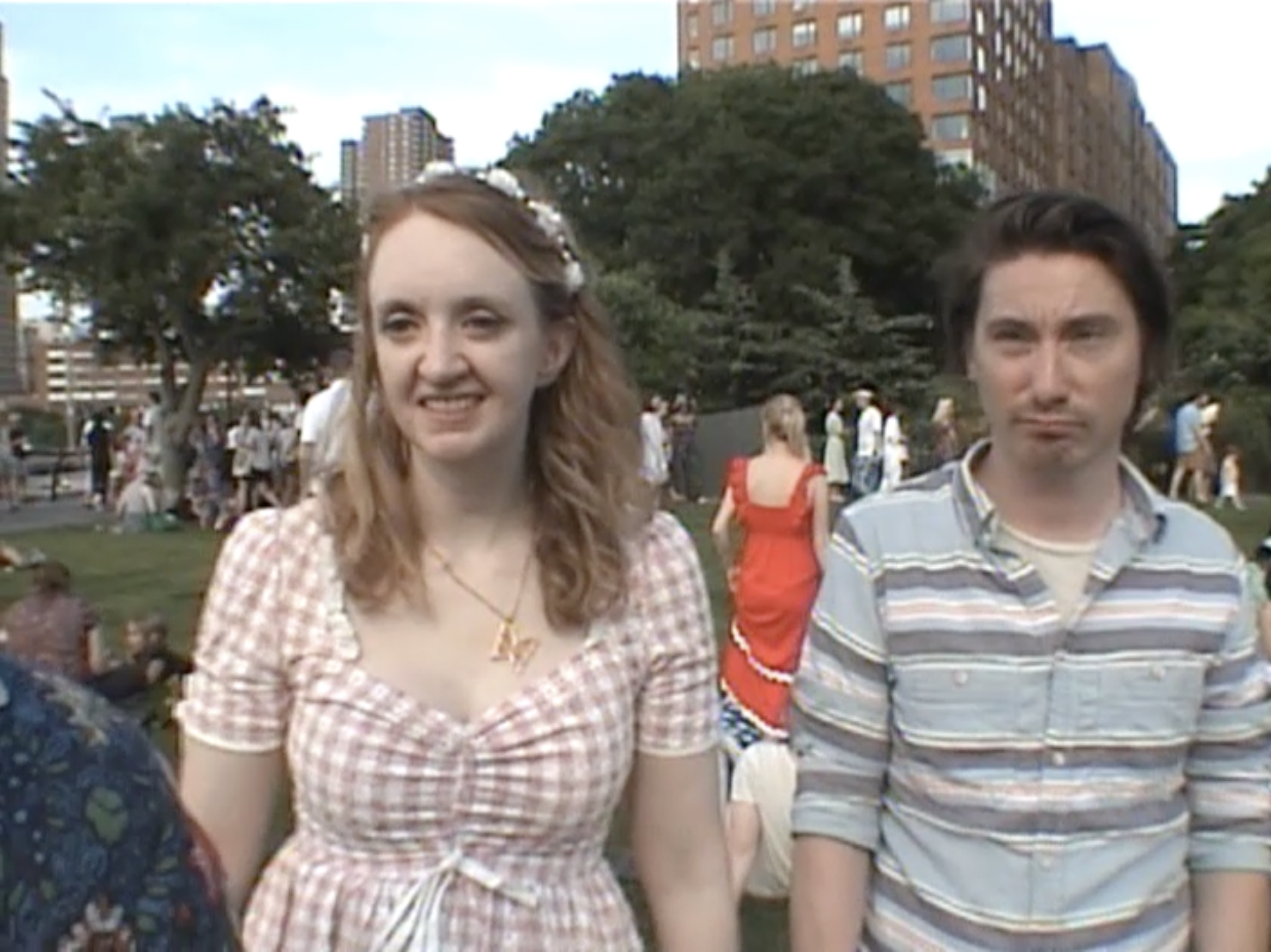 A young woman with long red hair wearing a gingham dress and a young man with dark hair wearing a striped shirt standing outside in a city park or plaza during daytime with tall buildings and other people in the background.
