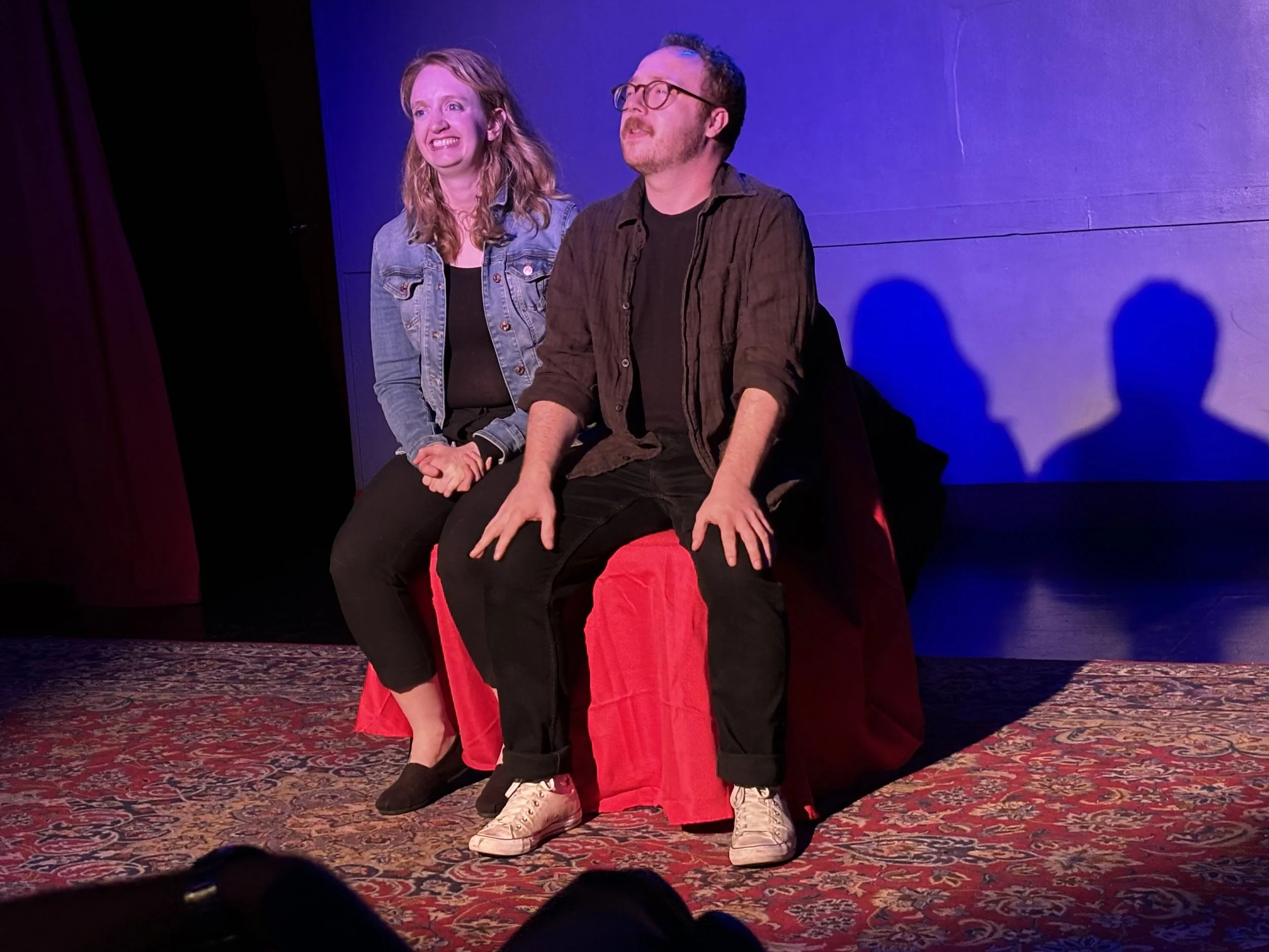 Two performers on stage, one woman and one man, sitting on a bench with a blue background and red patterned carpet, engaged in a performance or act.