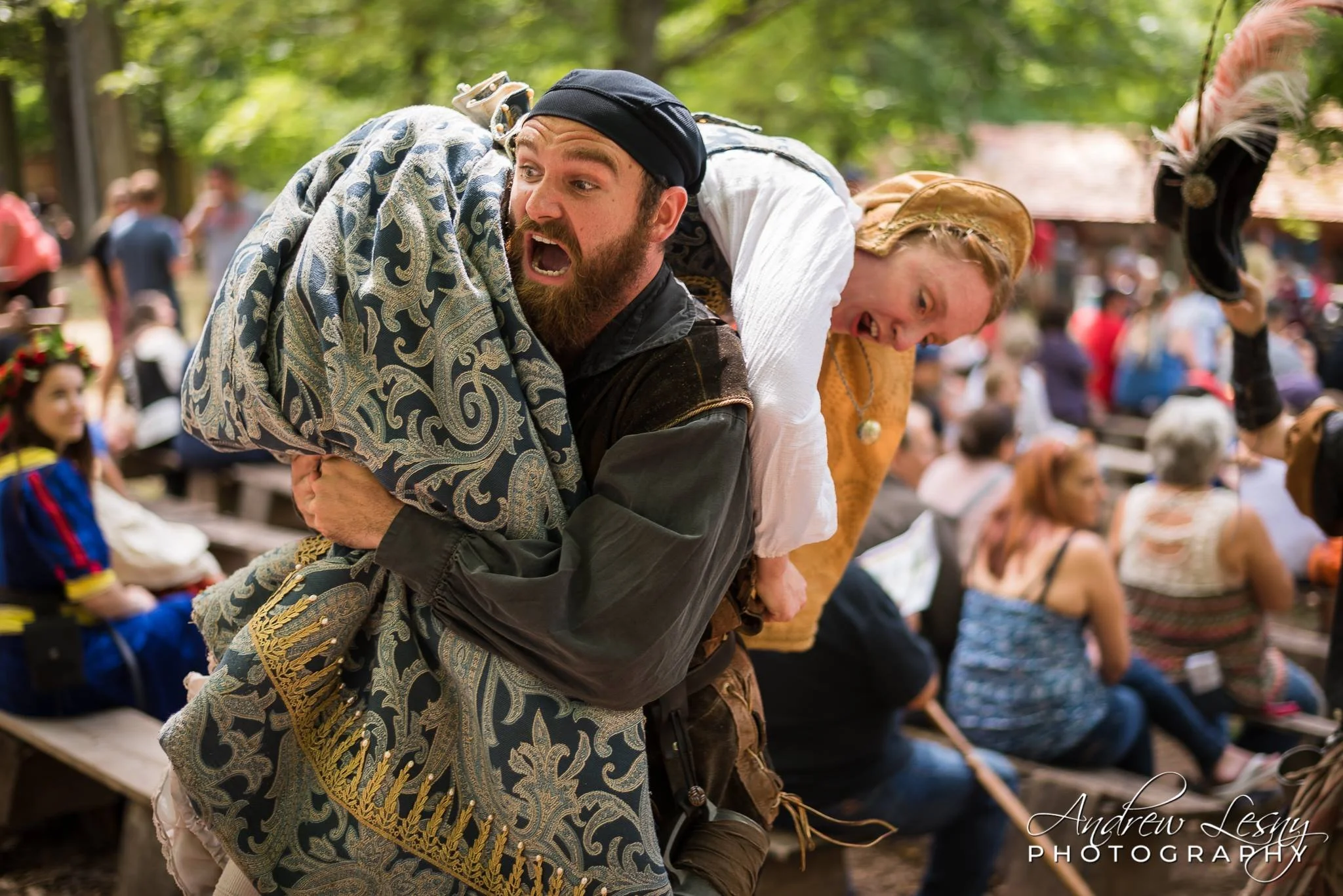 A man dressed in historical clothing participates in a reenactment or festival, carrying a woman on his back. The background shows people sitting on benches, some dressed in period costumes, with trees and a building, indicating an outdoor event.