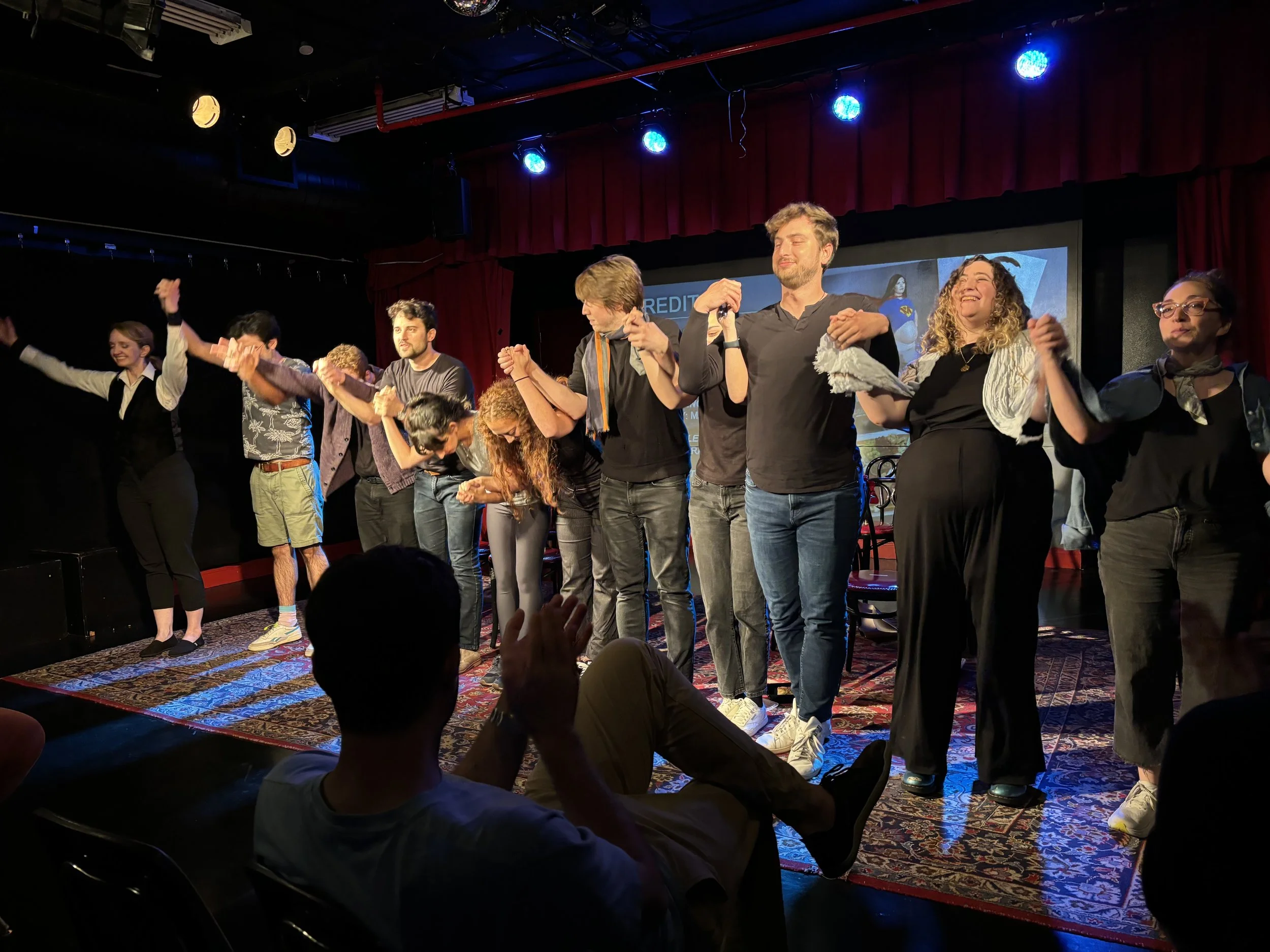 People standing on stage holding hands and bowing at the end of a performance or event in a dimly lit venue with red curtains and blue stage lights.
