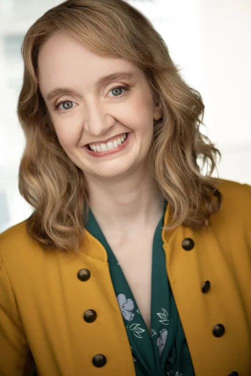 A smiling woman with blonde, wavy hair wearing a mustard yellow jacket with black buttons and a dark green top.