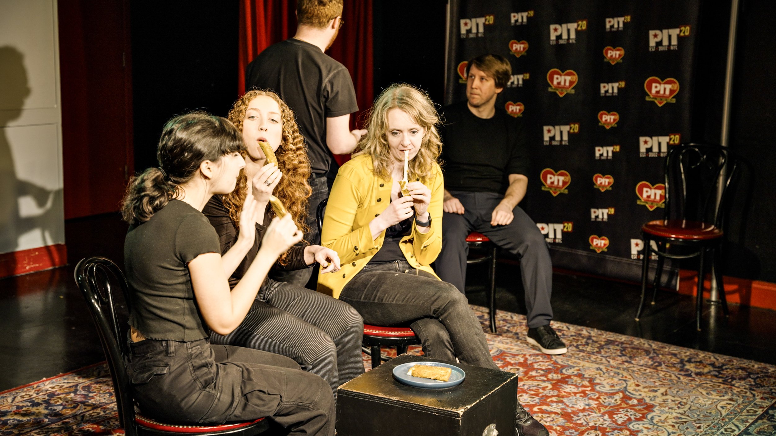 Group of five people sitting and standing on a stage with a Pittsburg-themed backdrop, eating and chatting with plates of food on the table.
