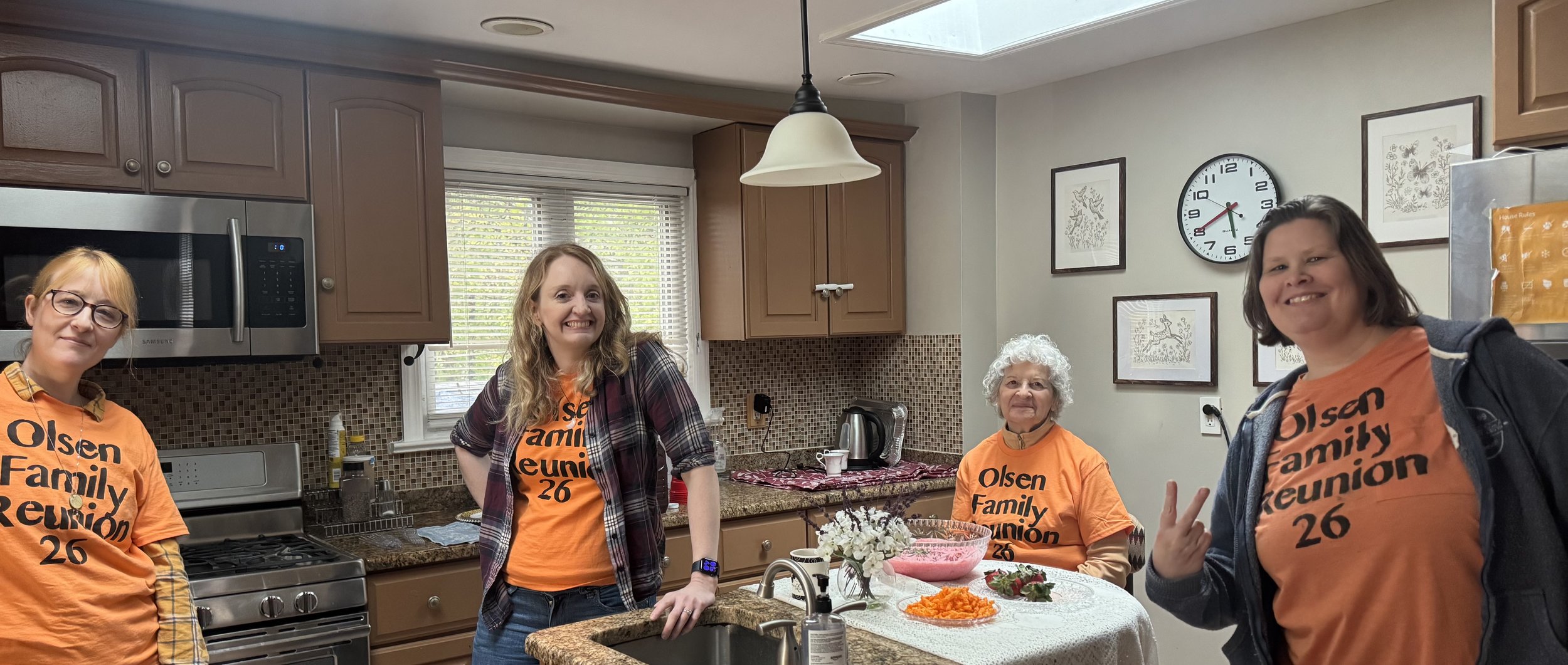 Four women standing in a kitchen, wearing matching orange shirts with 'Olsen Family Reuinion 26' printed on them, smiling at the camera during a family reunion event.
