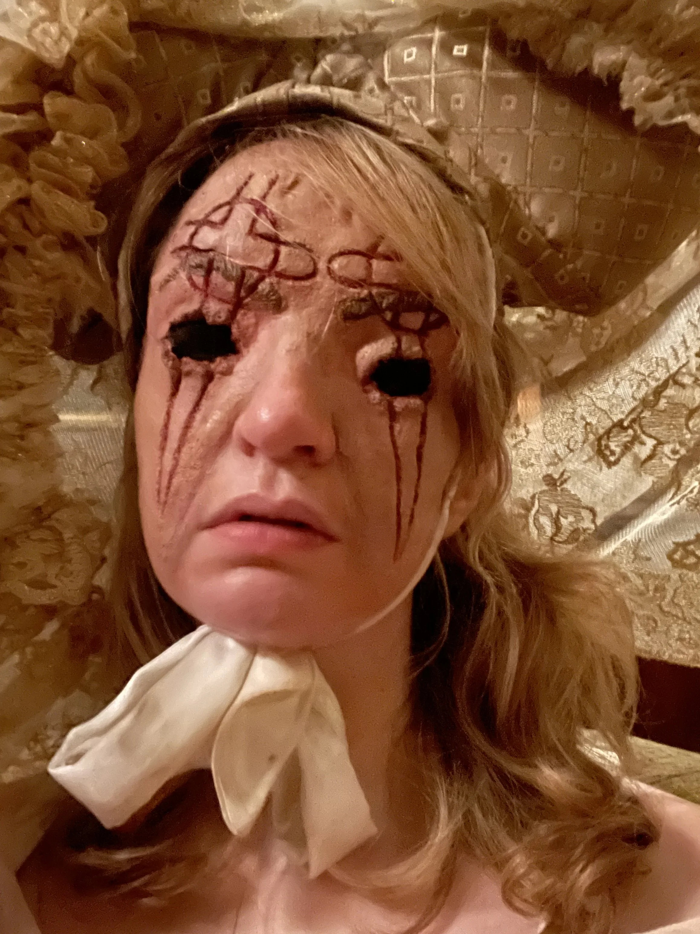 Close-up of a woman lying down, with makeup or special effects makeup on her face that resembles a zombie or monster, including dark spots around the eyes and streaks of red and black across her face.