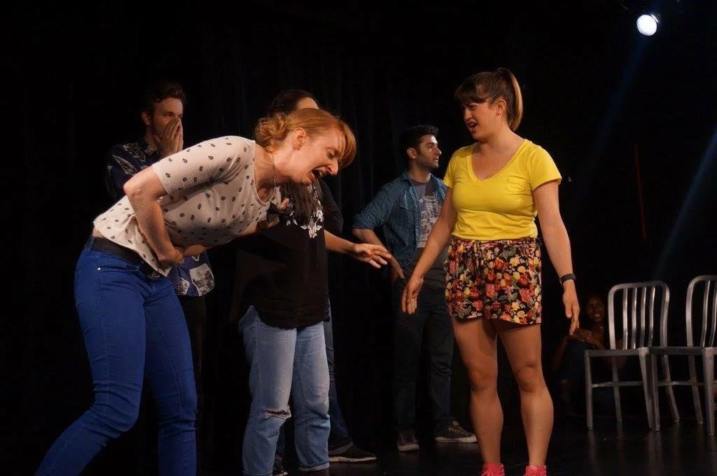 People performing comedy improv on stage, with three women in the foreground showing expressive reactions.
