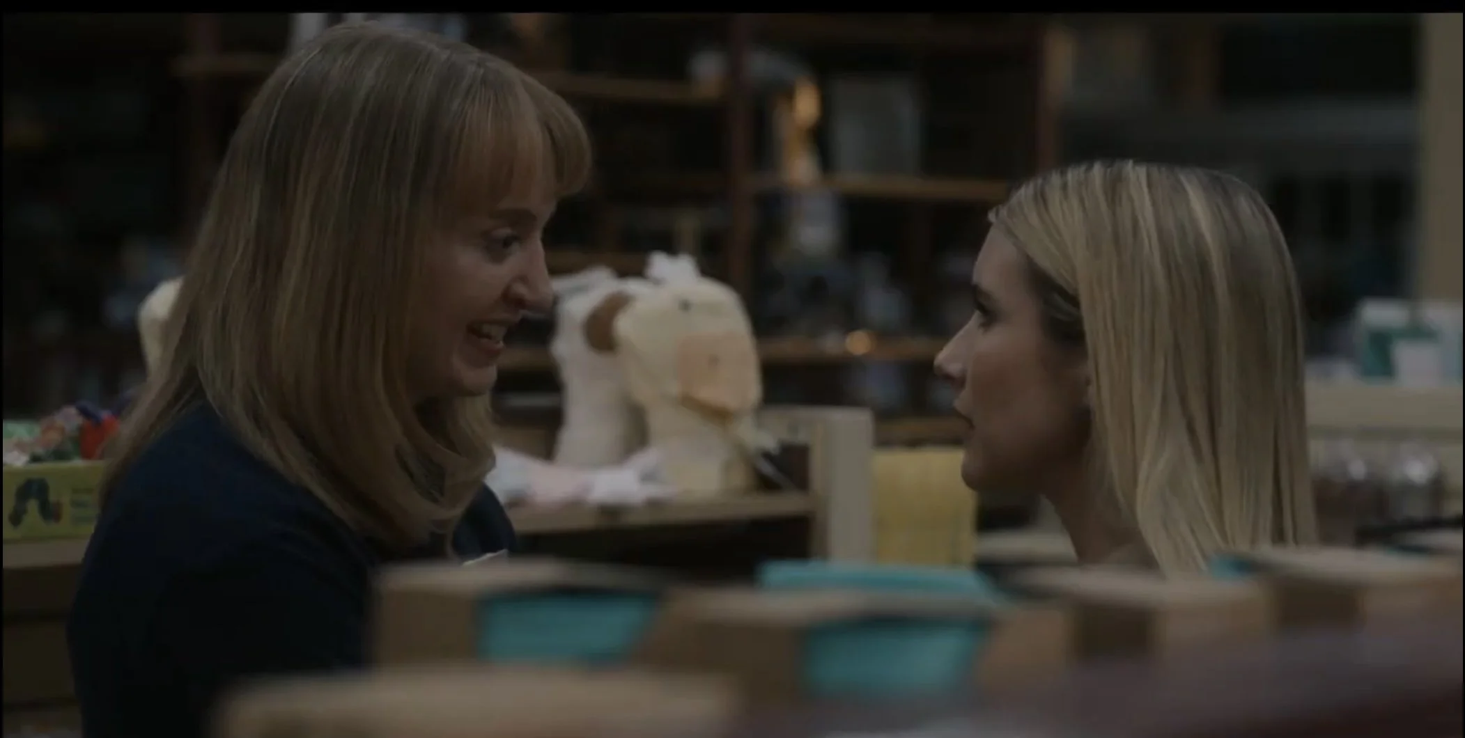 Two women face each other and smile in a store aisle, with shelves and various items in the background.