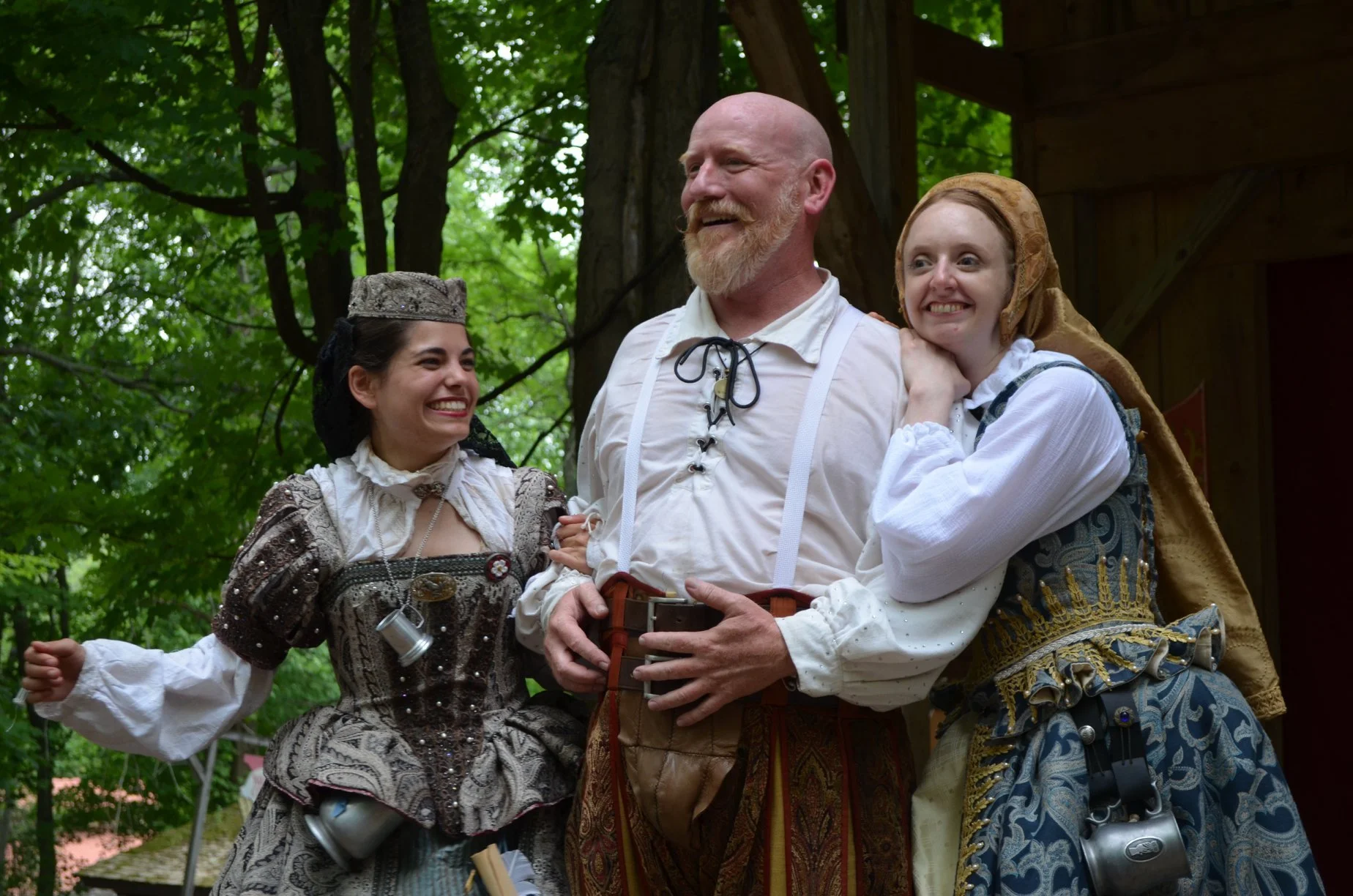 Three people dressed in historic or renaissance costumes standing outdoors among green trees, smiling and posing happily for a photo.