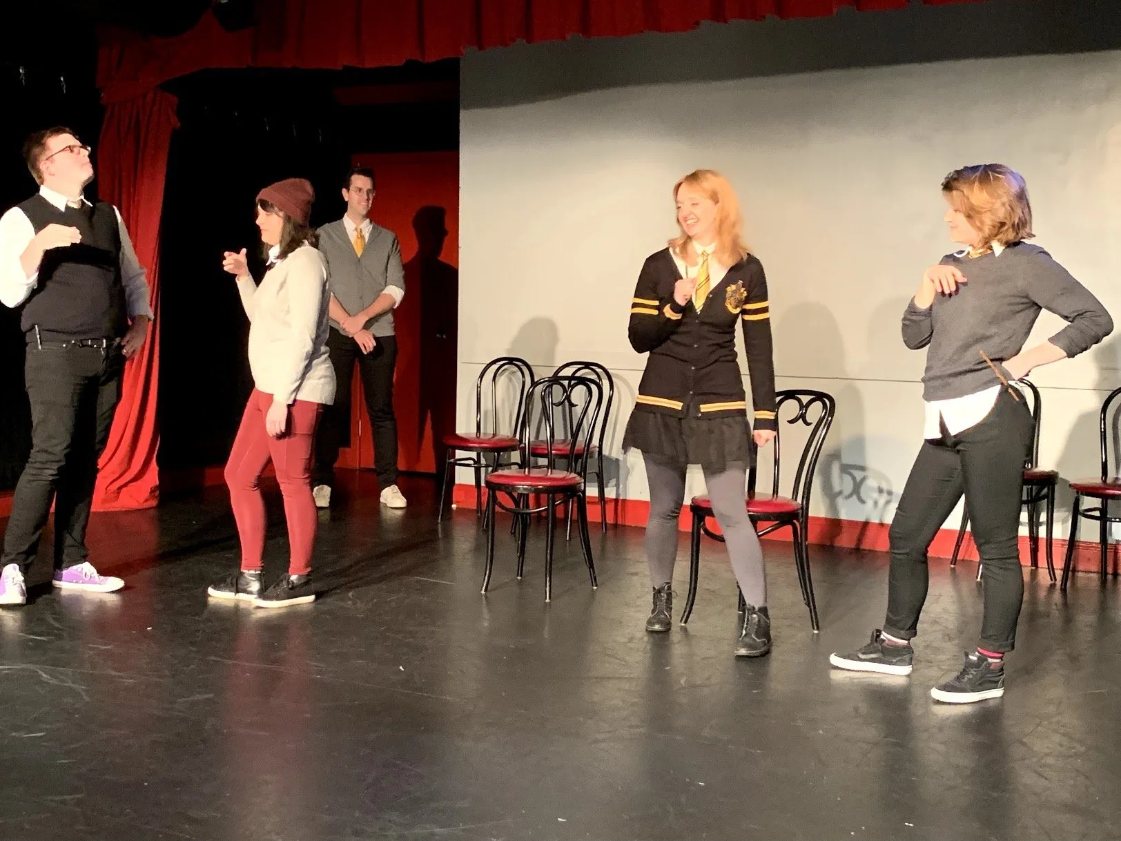 Group of five people on a stage with black chairs and red curtains, participating in a theatrical performance or rehearsal, some standing and talking, others observing.