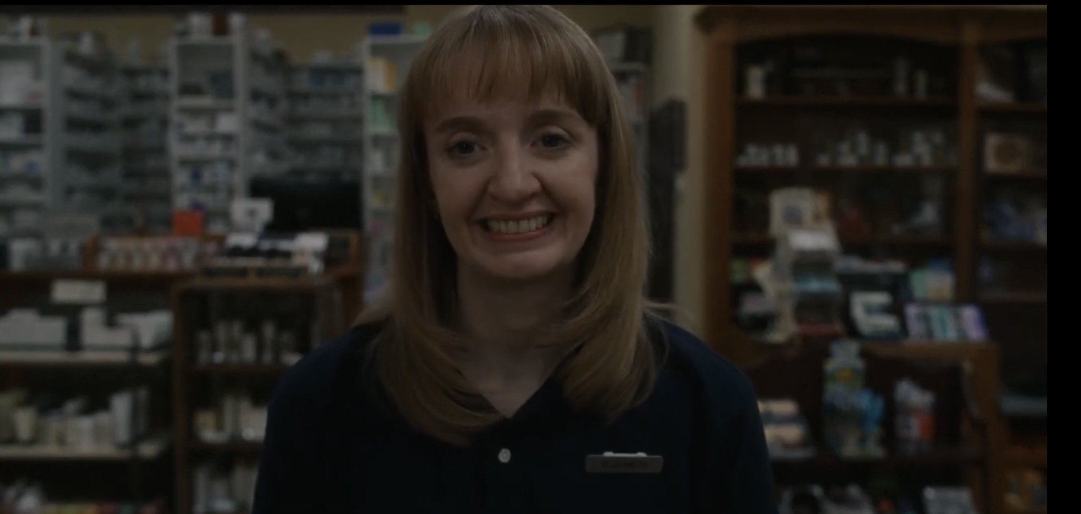 A woman with shoulder-length brown hair and bangs smiling at the camera, in a bookstore or library with shelves filled with books in the background.