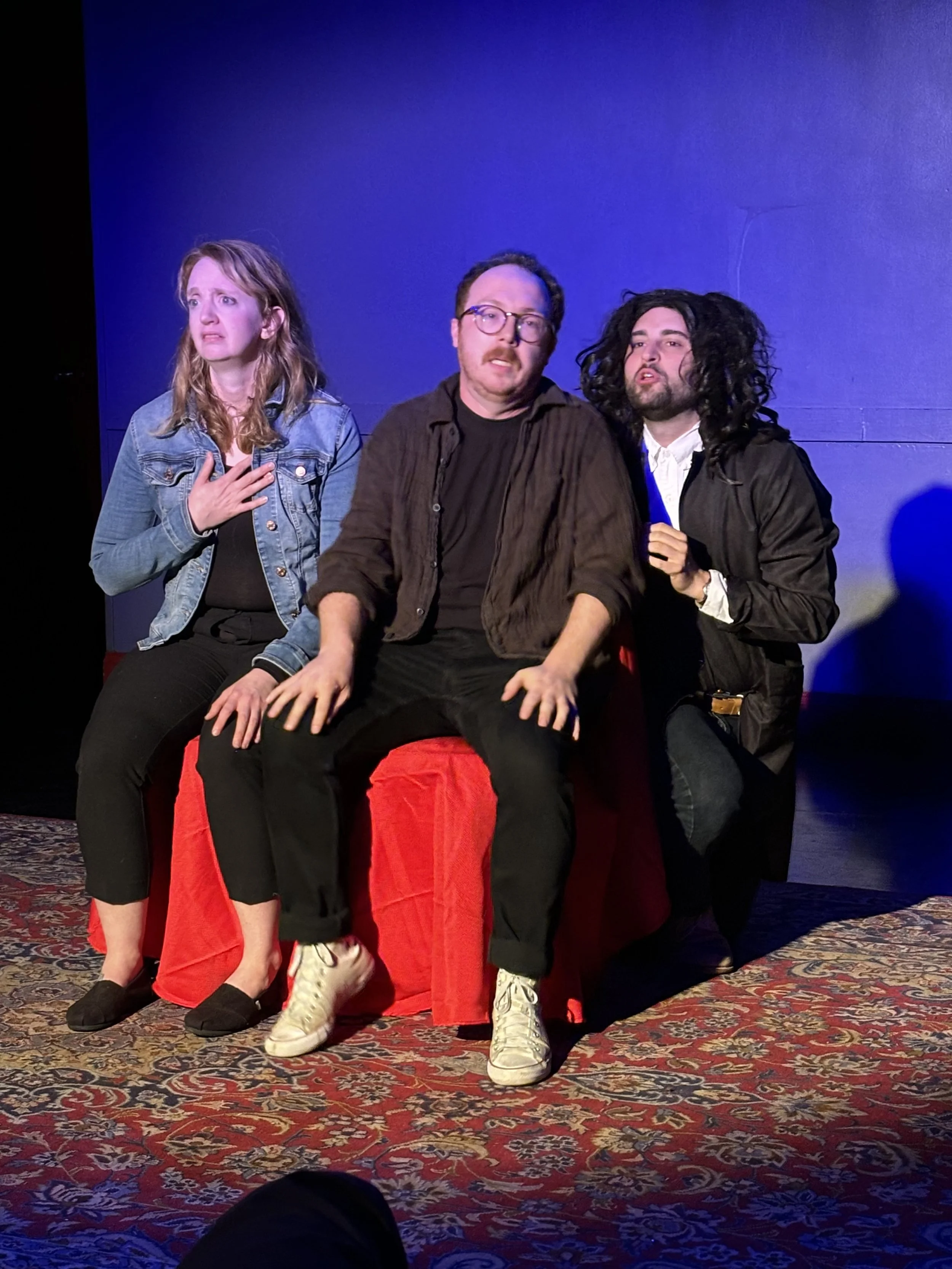 Three actors on a stage with a blue background. The woman on the left has light brown hair, wearing a denim jacket and black pants, with a surprised expression and hand on chest. The man in the middle has glasses, brown hair, wearing a brown shirt, s