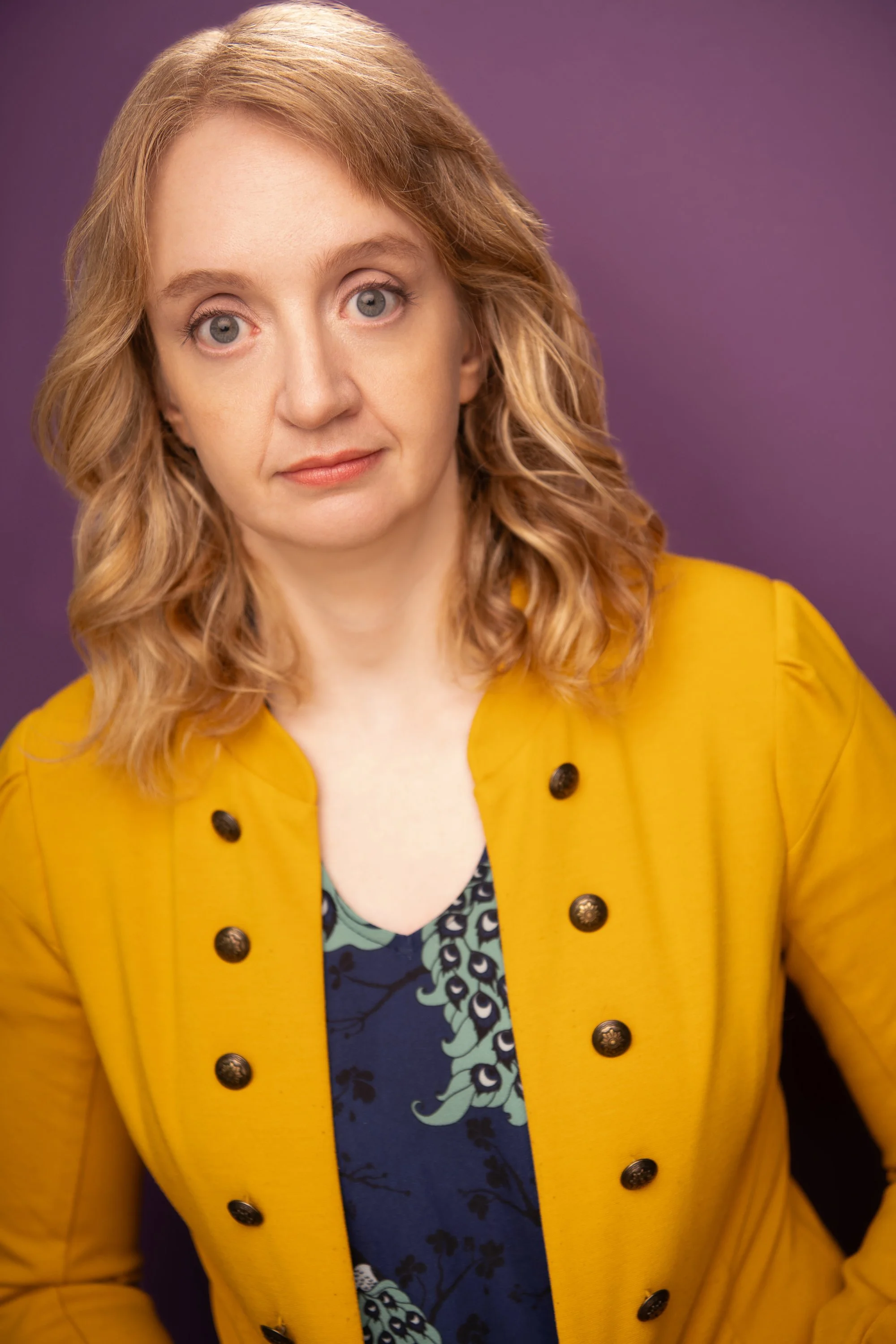 Close-up of a woman with red hair wearing a mustard yellow coat and a patterned top, against a purple background.