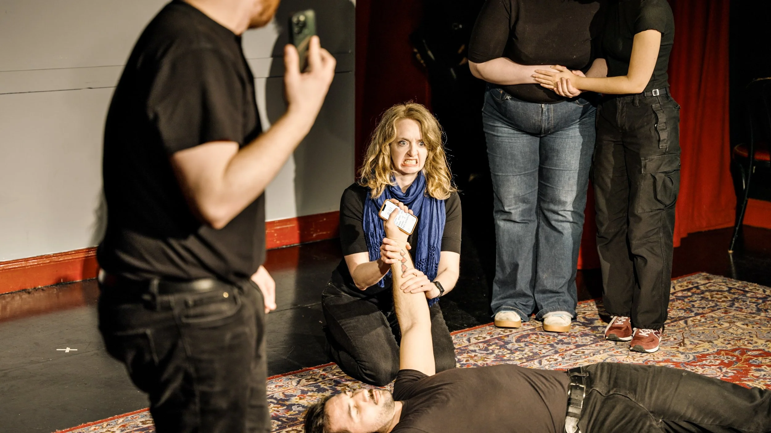 A woman with curly blonde hair sitting on the floor with a distressed expression, clutching a small dog. Several other people stand around her, some with concerned expressions, in a dimly lit room with a patterned rug.