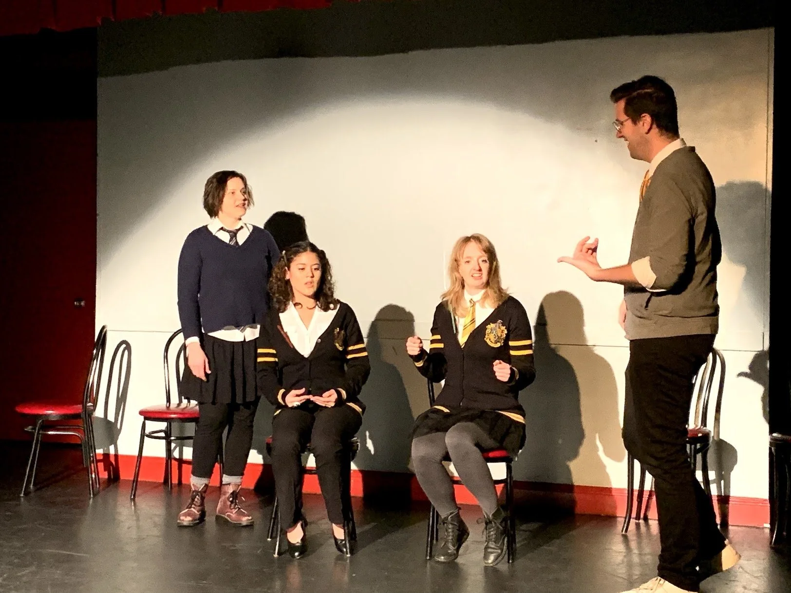 Four young women dressed in Harry Potter-themed uniforms and one man in a gray suit with glasses on a stage, with the man appearing to be performing or speaking to the women. The stage has black chairs with red seats, and a plain backdrop.