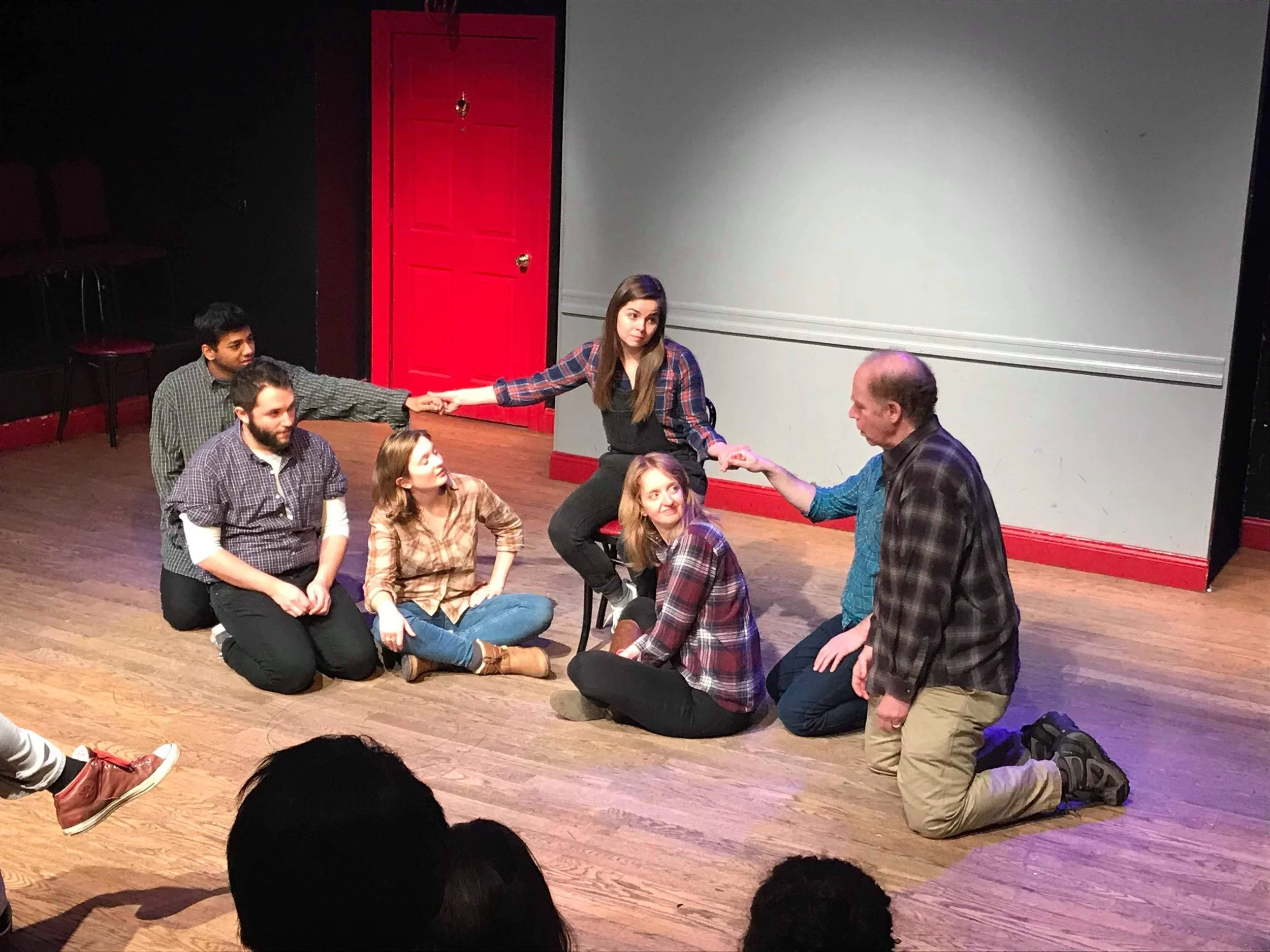 A group of people participating in a theatrical or improv performance on a wooden stage. Some individuals are sitting on the floor, others kneeling or standing, with two people holding hands and others appearing to be engaged in the scene. There is a