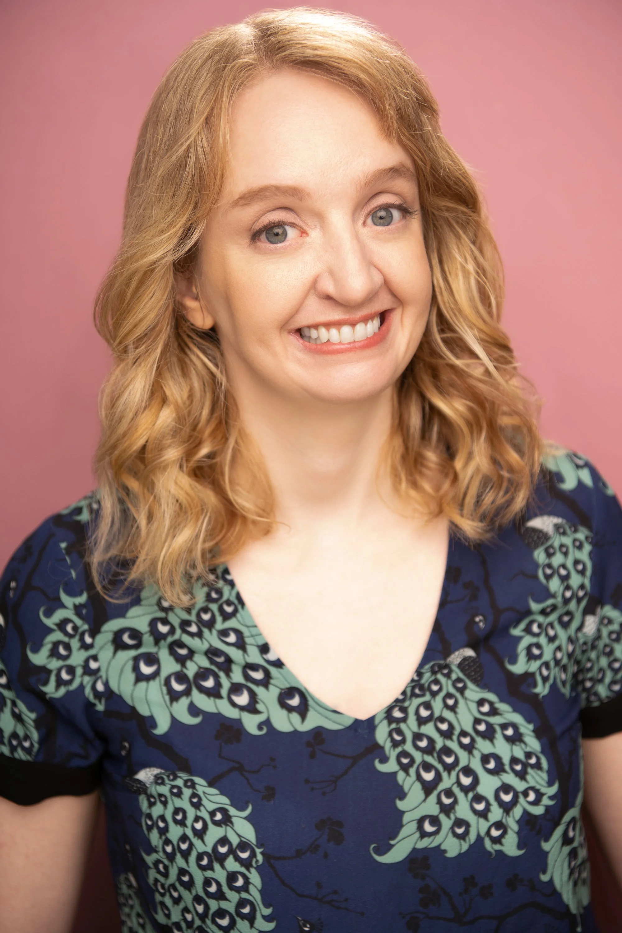 Close-up photograph of a young woman with curly, blonde hair, smiling, wearing a peacock feather pattern dress with a pink background.