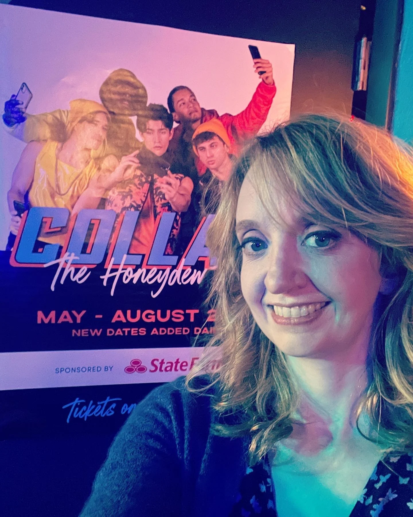 A woman with curly red hair smiling in front of a promotional poster for the musical 'Cursed: The Homestead' showing a group of young people taking selfies.