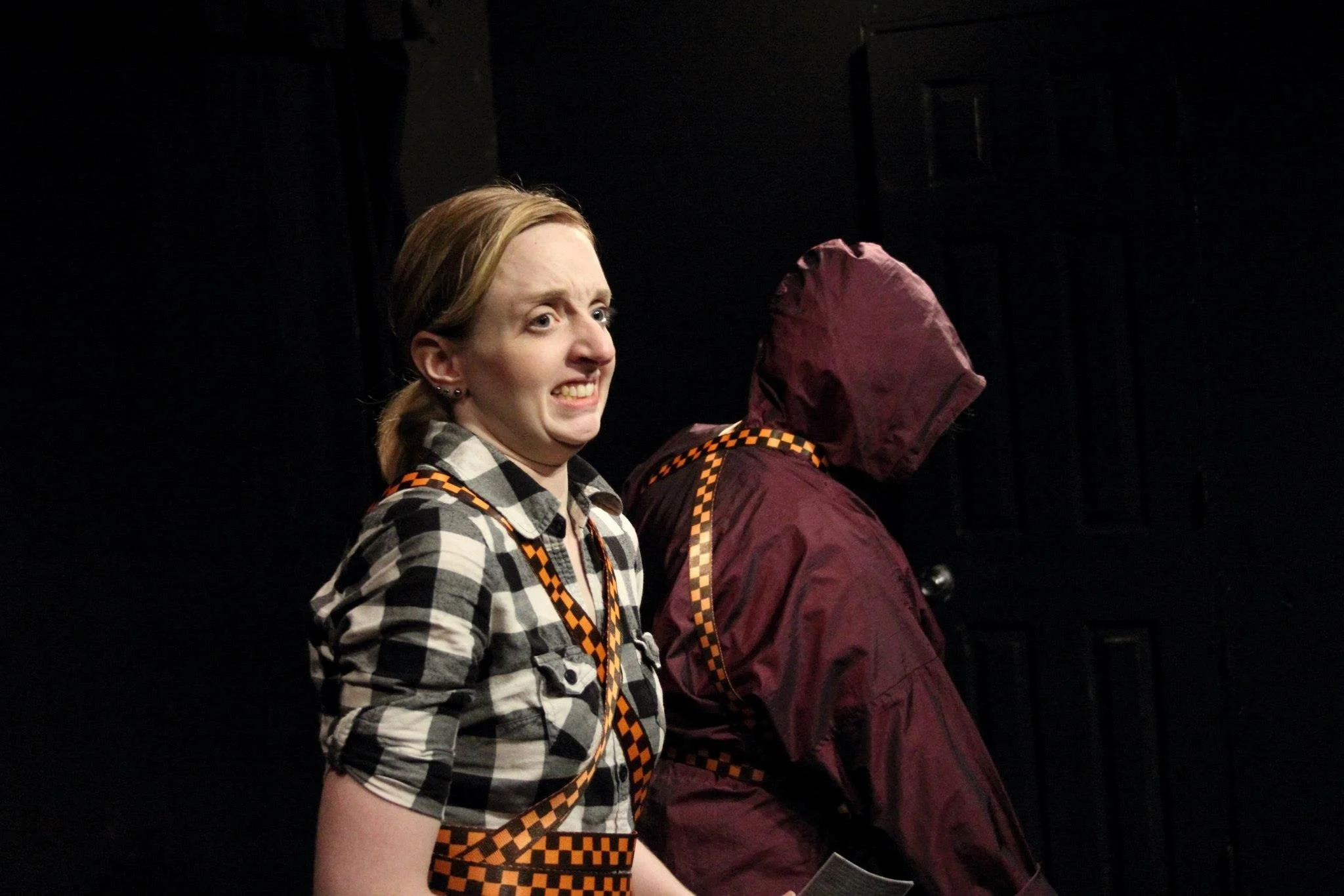 A woman with light skin and blonde hair in a ponytail, wearing a black and white checkered shirt, making a funny face with her eyes wide and teeth showing. She has a checkered lanyard around her neck. Next to her is a person in a maroon jacket with a