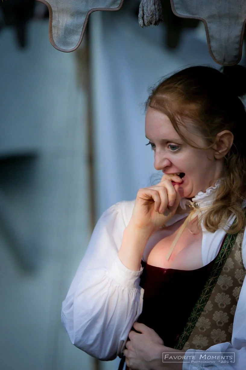 A woman dressed in historical or fantasy clothing, with a white blouse and a patterned vest, is smiling and biting her finger while looking down.