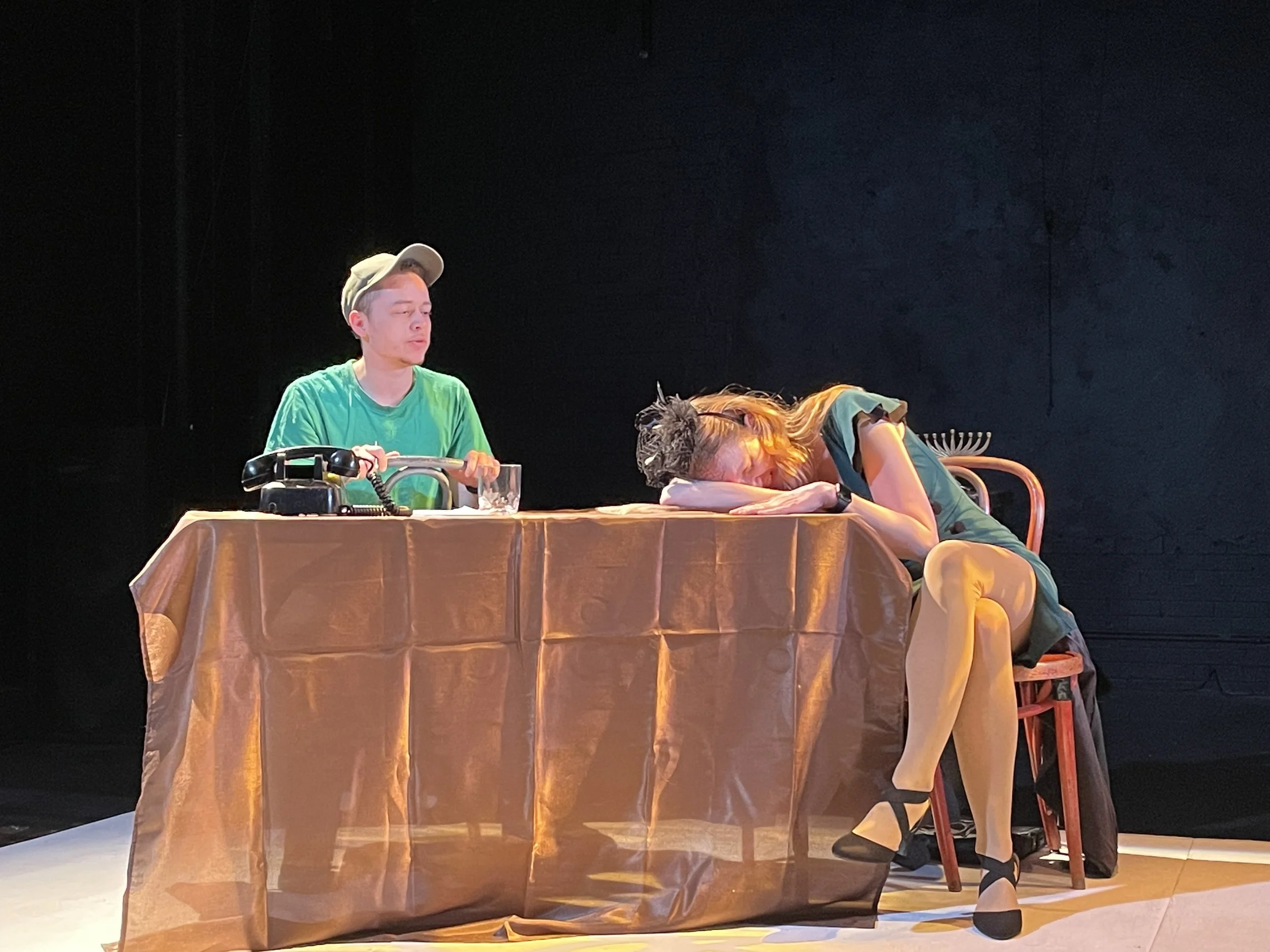 Two people sitting at a table with a gold-colored tablecloth, against a black background. One person is sitting upright wearing a green shirt and beige cap, and the other person, with long hair, is resting their head and arms on the table, wearing a 