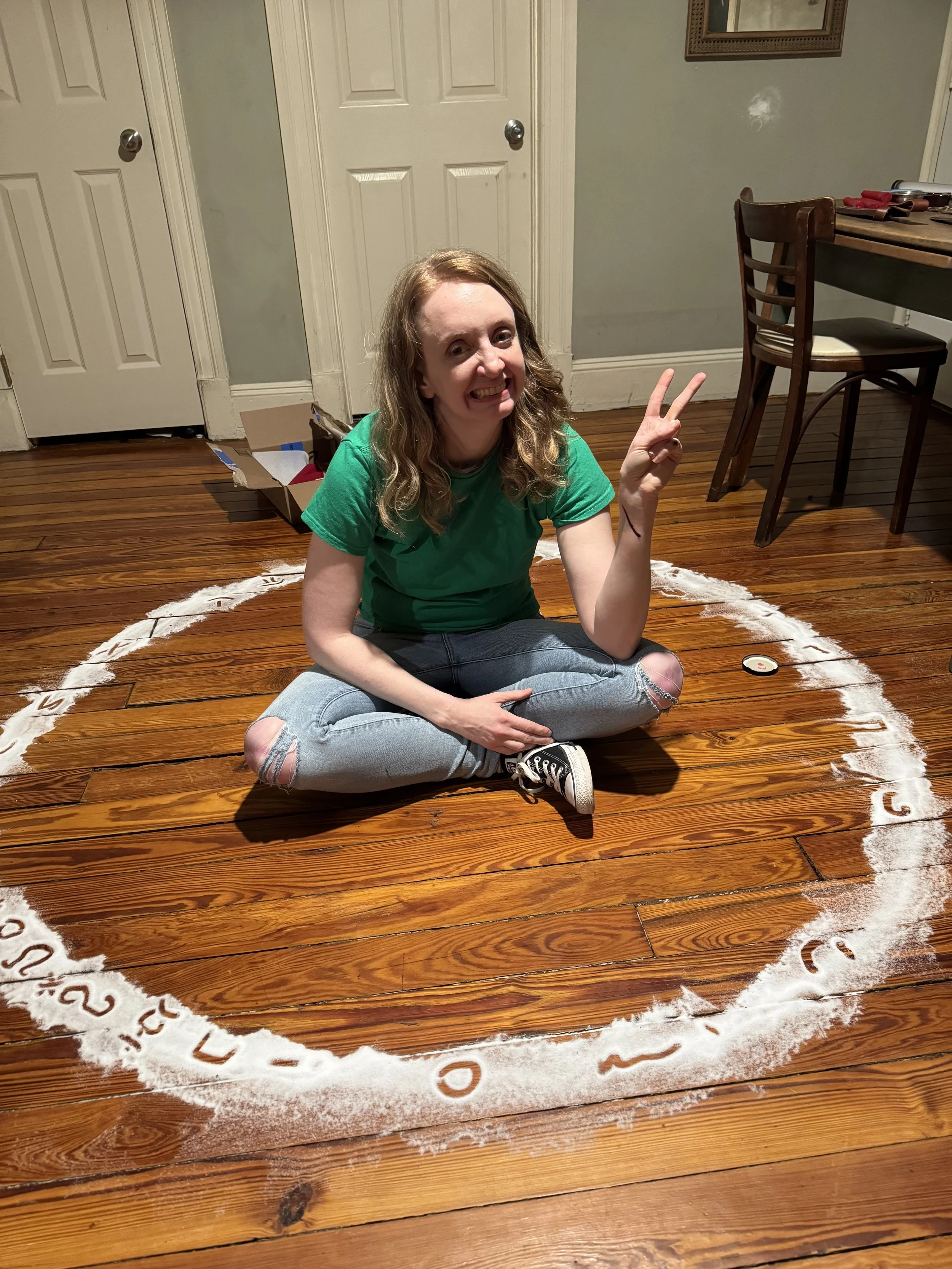 A woman sitting on a wooden floor inside a circle drawn in white powder, making a peace sign with her right hand and smiling.