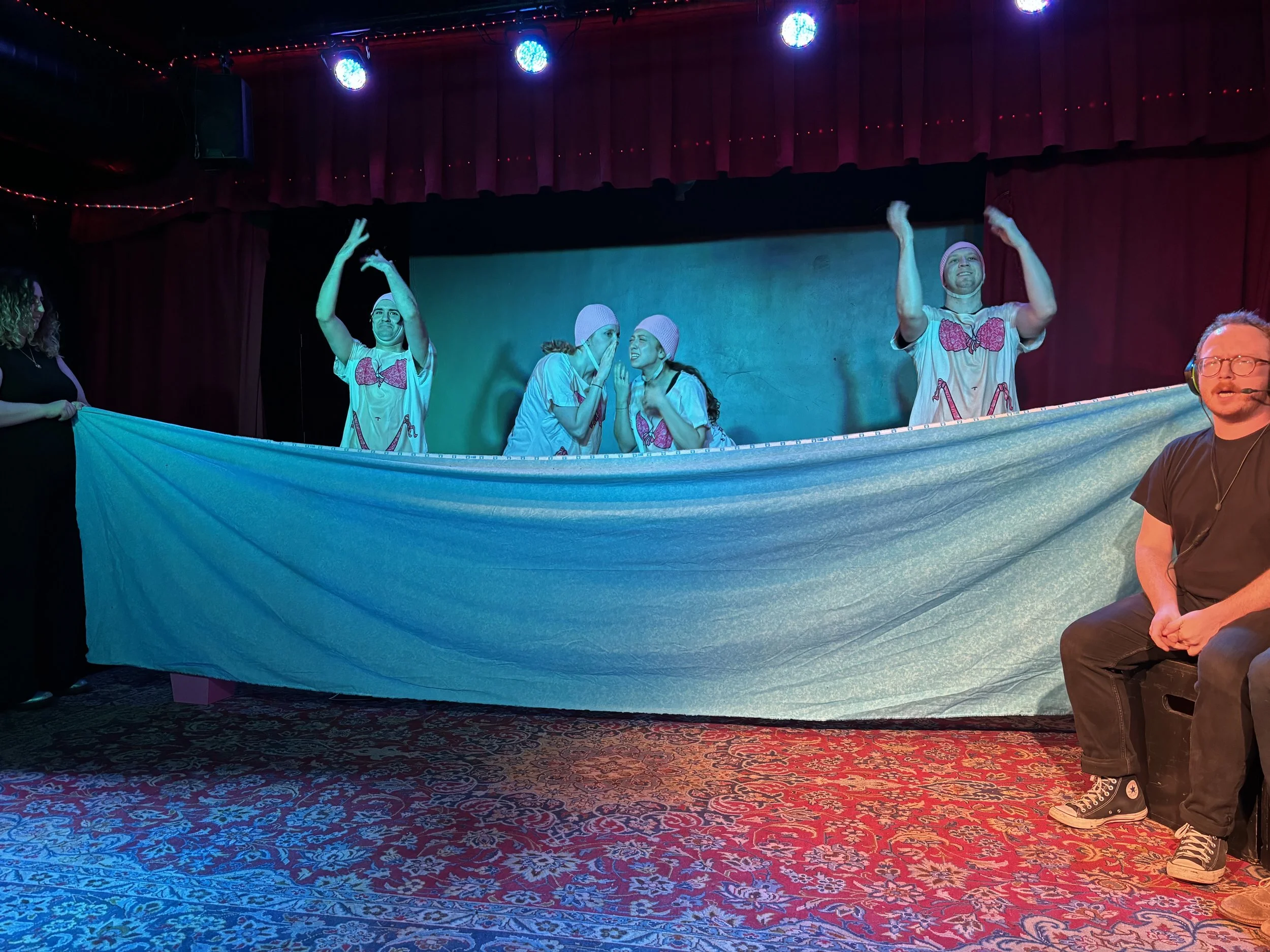 People dressed in pajamas with pink bows perform on a stage with a long blue cloth and a red curtain, under colorful stage lighting.