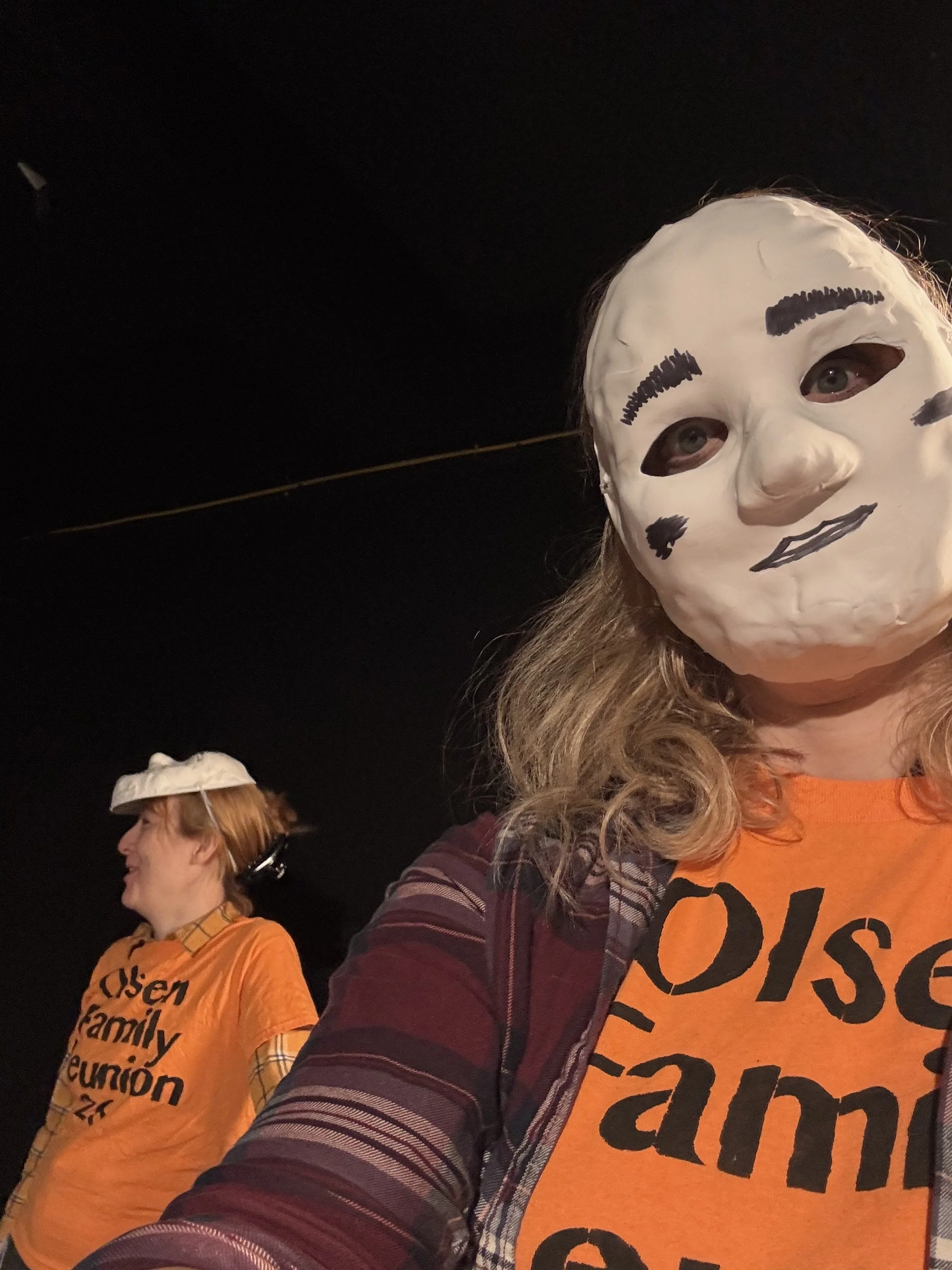 Two women wearing masks and orange shirts, possibly participating in a campaign or event, standing under a dark sky at night.